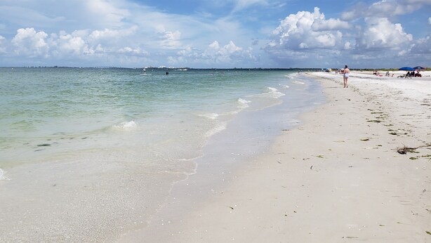 Una playa con aguas cristalinas, orilla arenosa y el horizonte de una ciudad a lo lejos.
