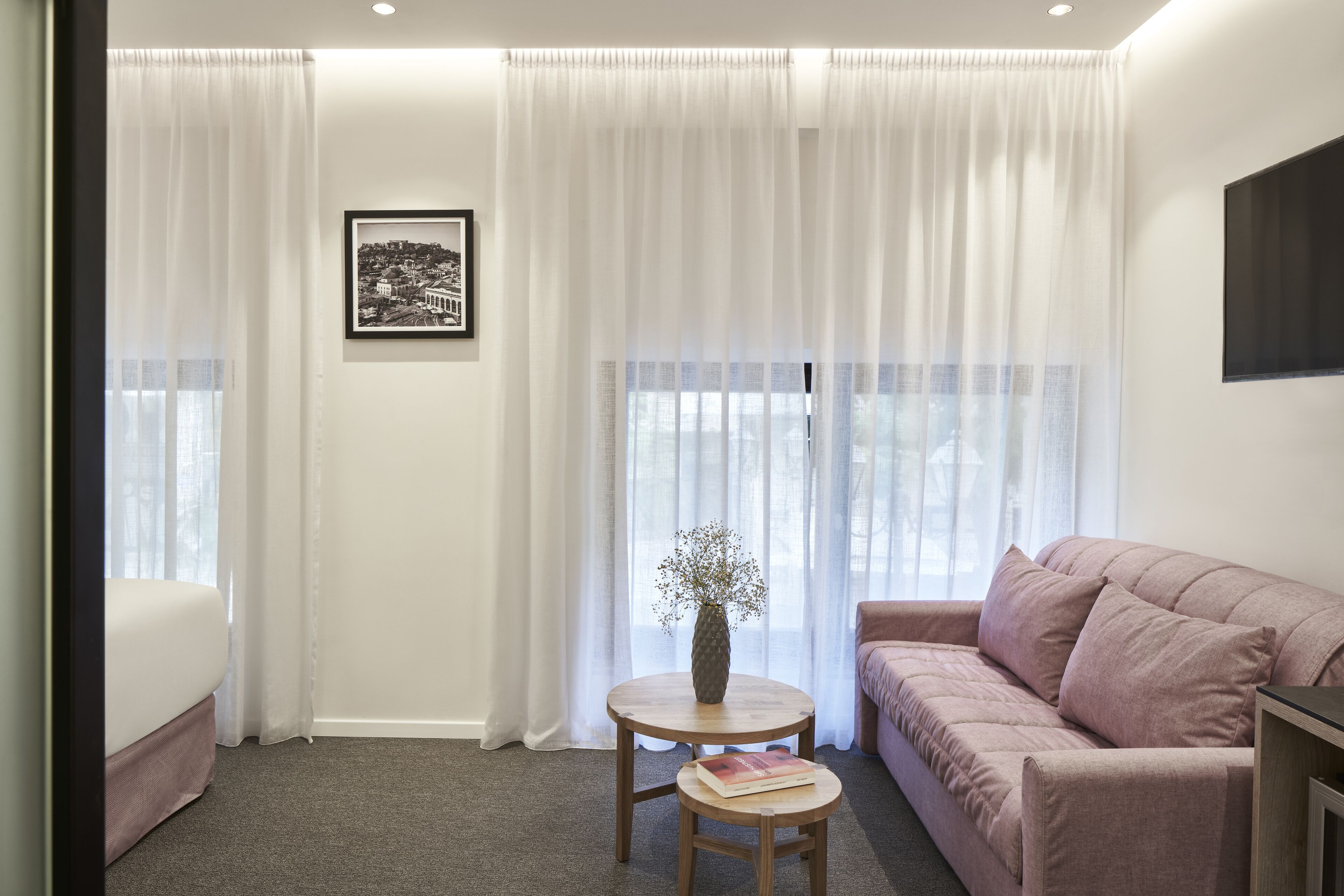 A modern hotel room with a bed, a pink sofa, a small round table with a vase, and a framed picture on the wall.