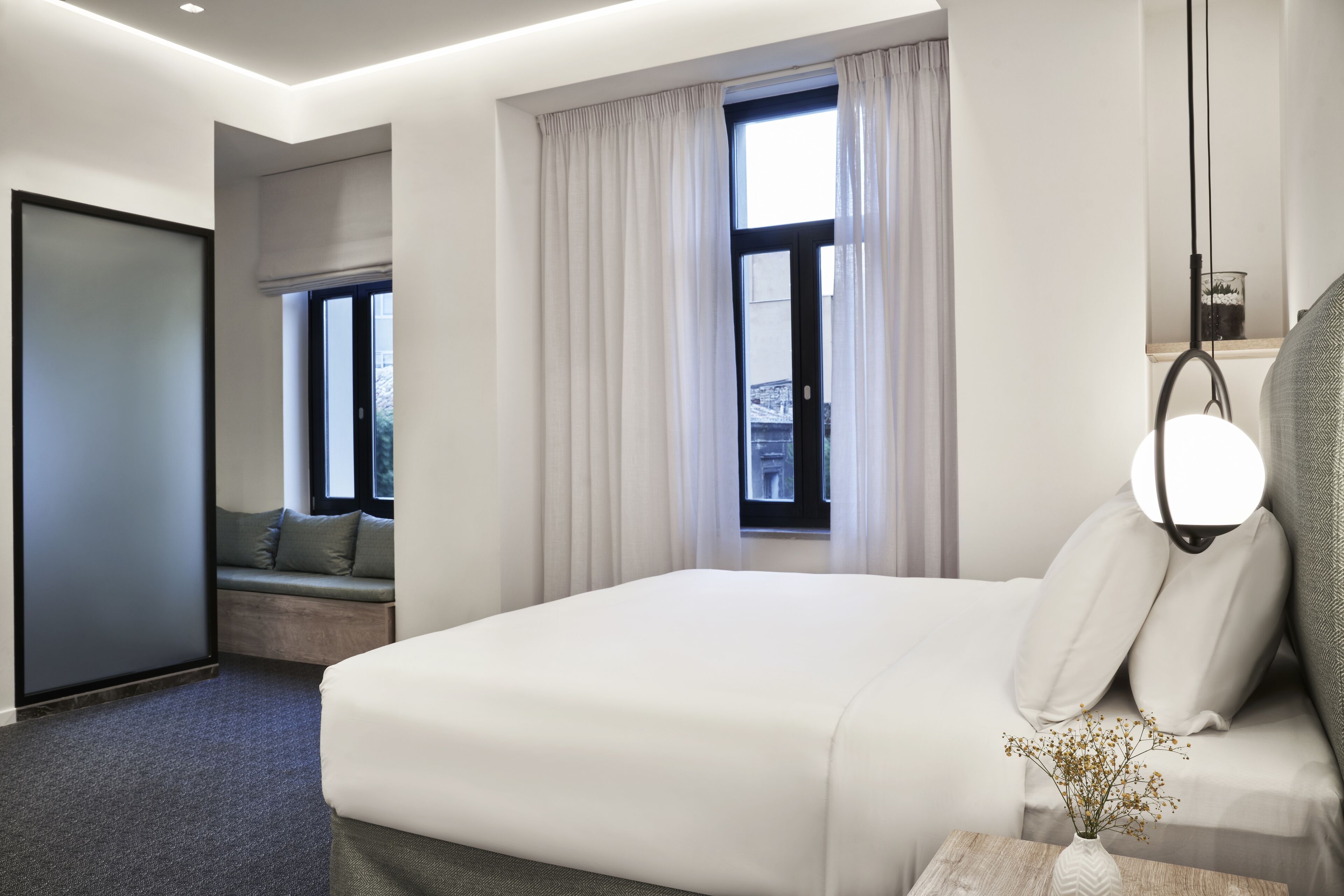 A modern hotel room with a large bed, a bedside table with a lamp, a window with curtains, and a view of the city.