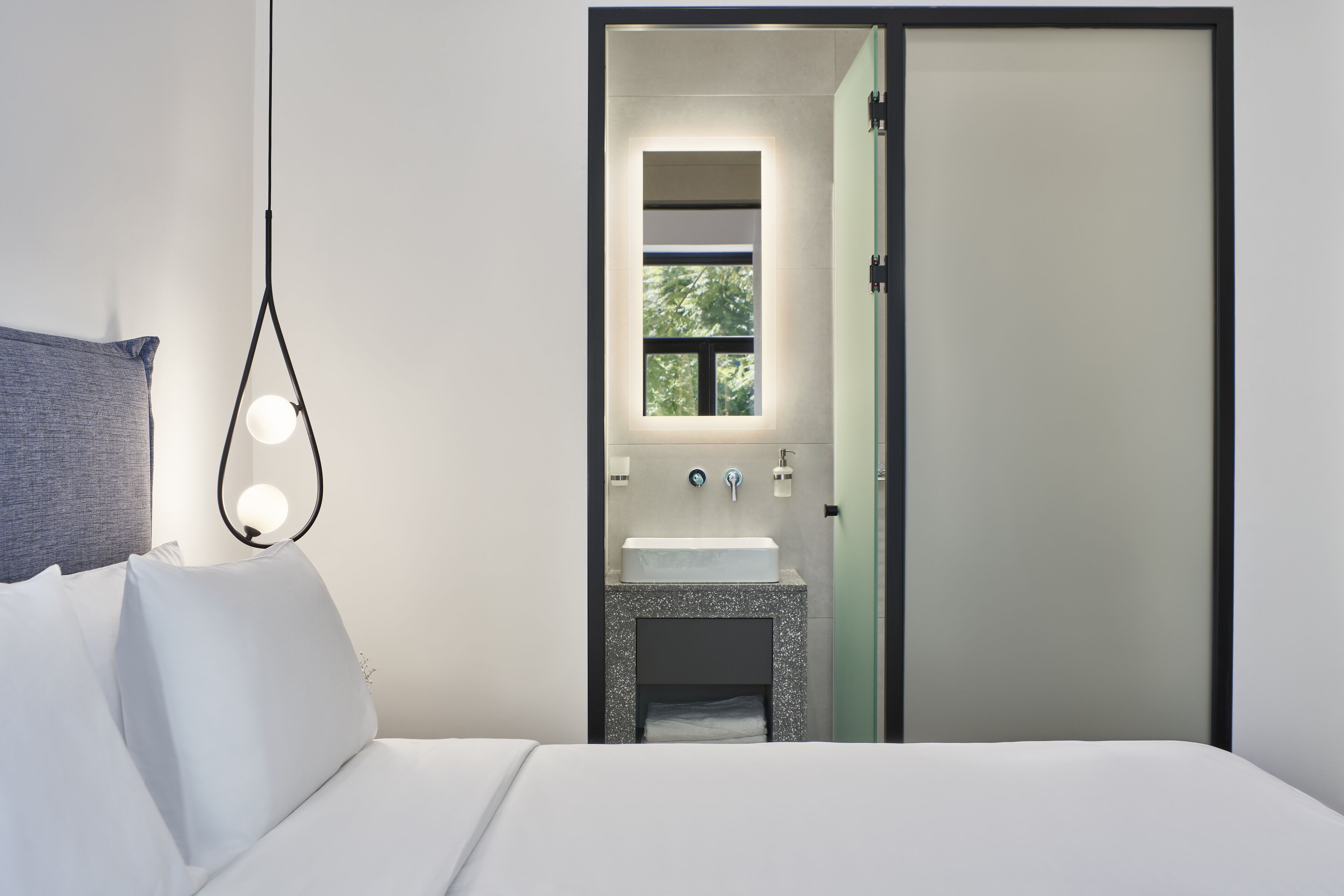 Een moderne hotelkamer met een bed, een bedlampje, een raam en een badkamer door een open deur zichtbaar.