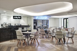 Um lobby moderno com uma placa indicando 'Lobby Bar', uma máquina de venda automática de comida e uma área de estar com mesas e cadeiras.