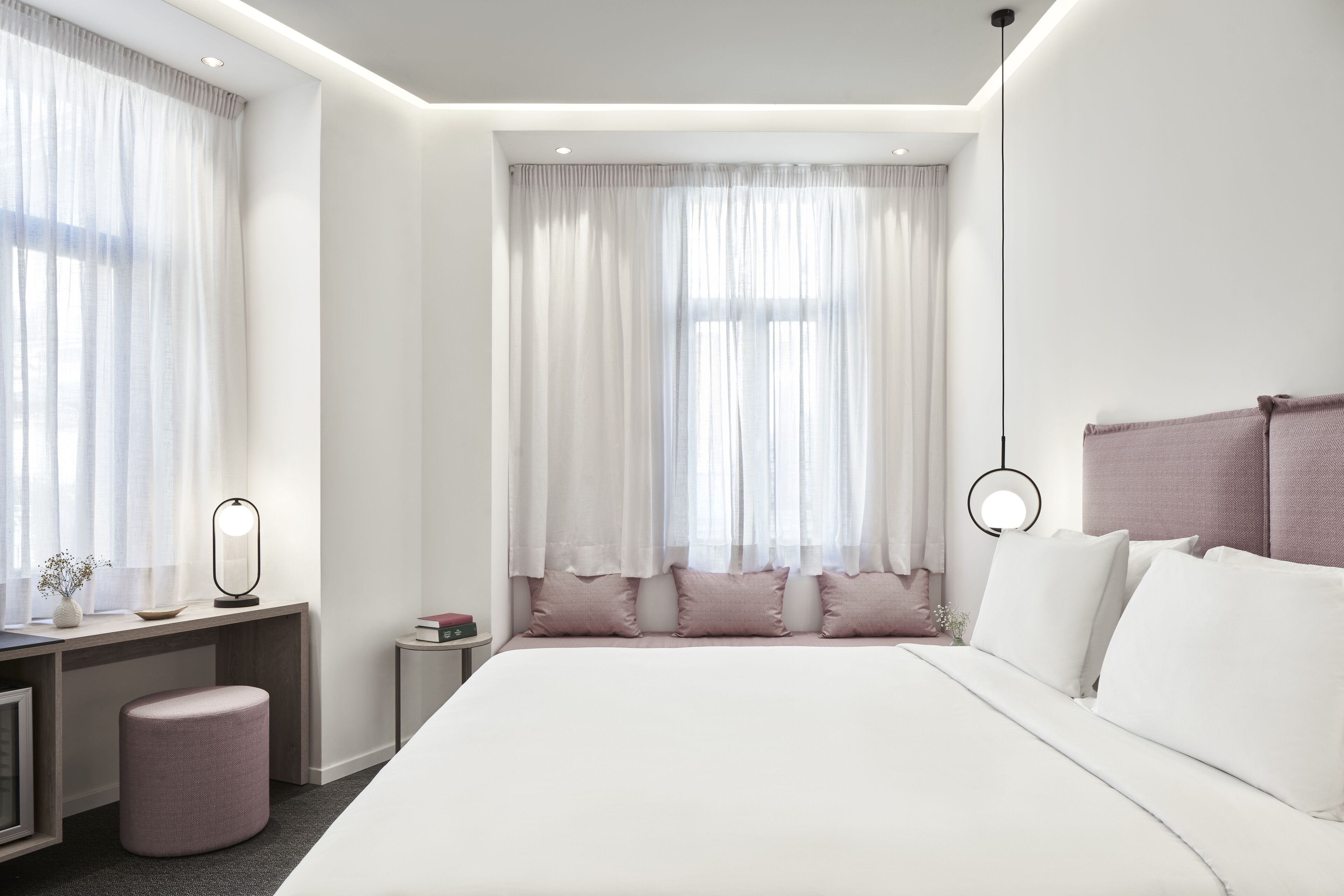 Una camera d'hotel moderna con un letto grande, una scrivania con lampada e una finestra con tende leggere.
