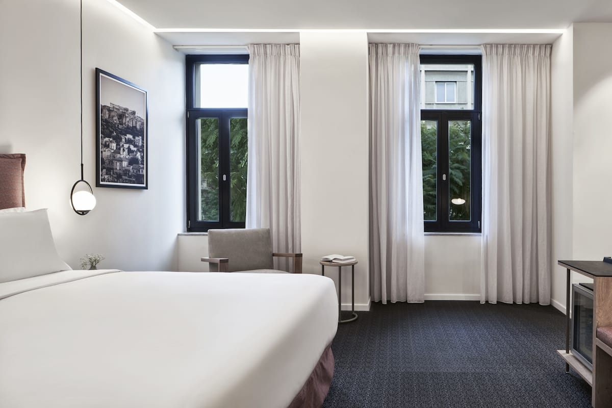 Habitación de hotel con una cama grande, una silla, una mesita y un televisor. Hay dos ventanas con cortinas, y un cuadro en la pared.