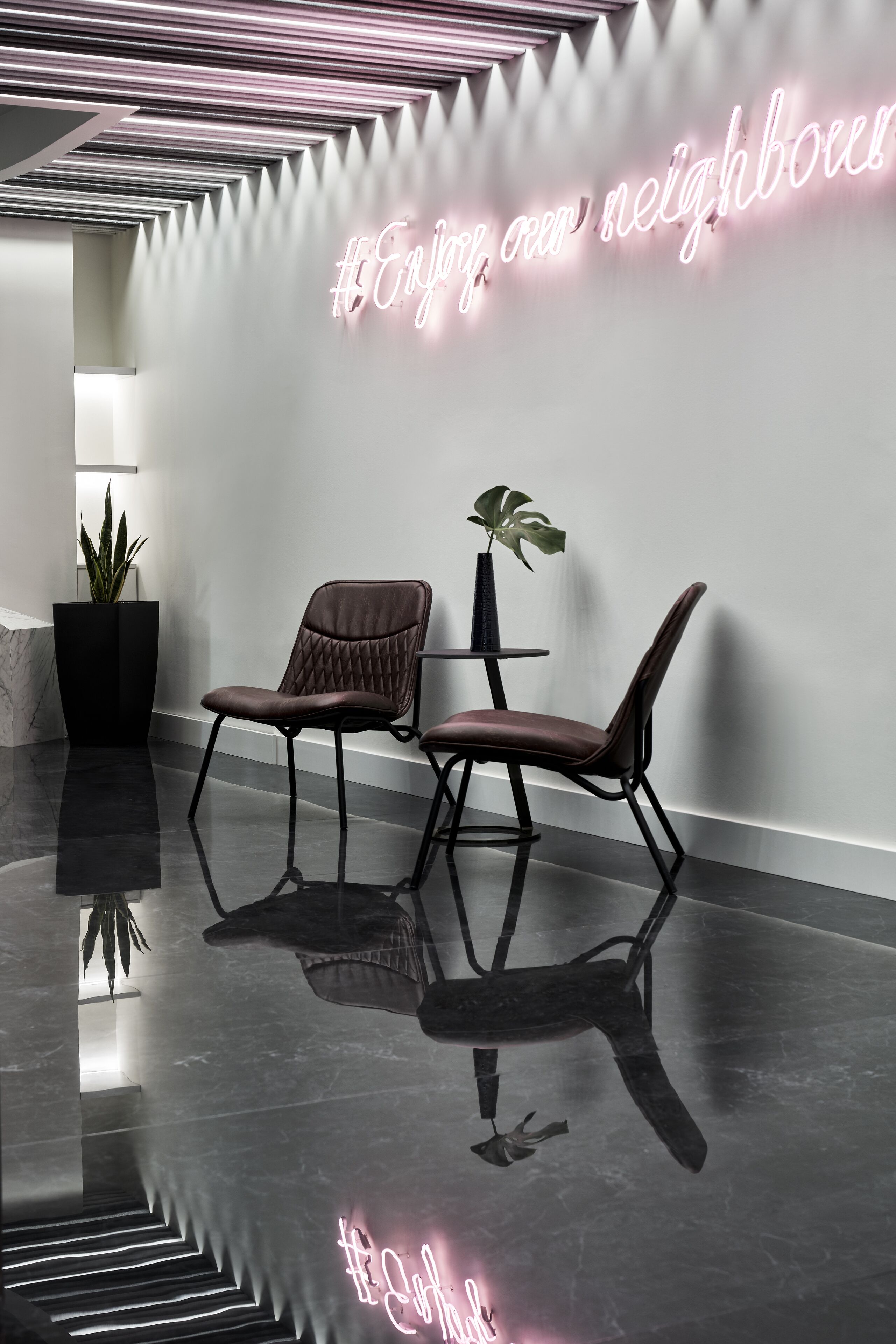 Un hall d'entrée moderne avec un panneau néon sur lequel on peut lire « #Profitez de nos voisins » et deux fauteuils ornés de plantes en pot.