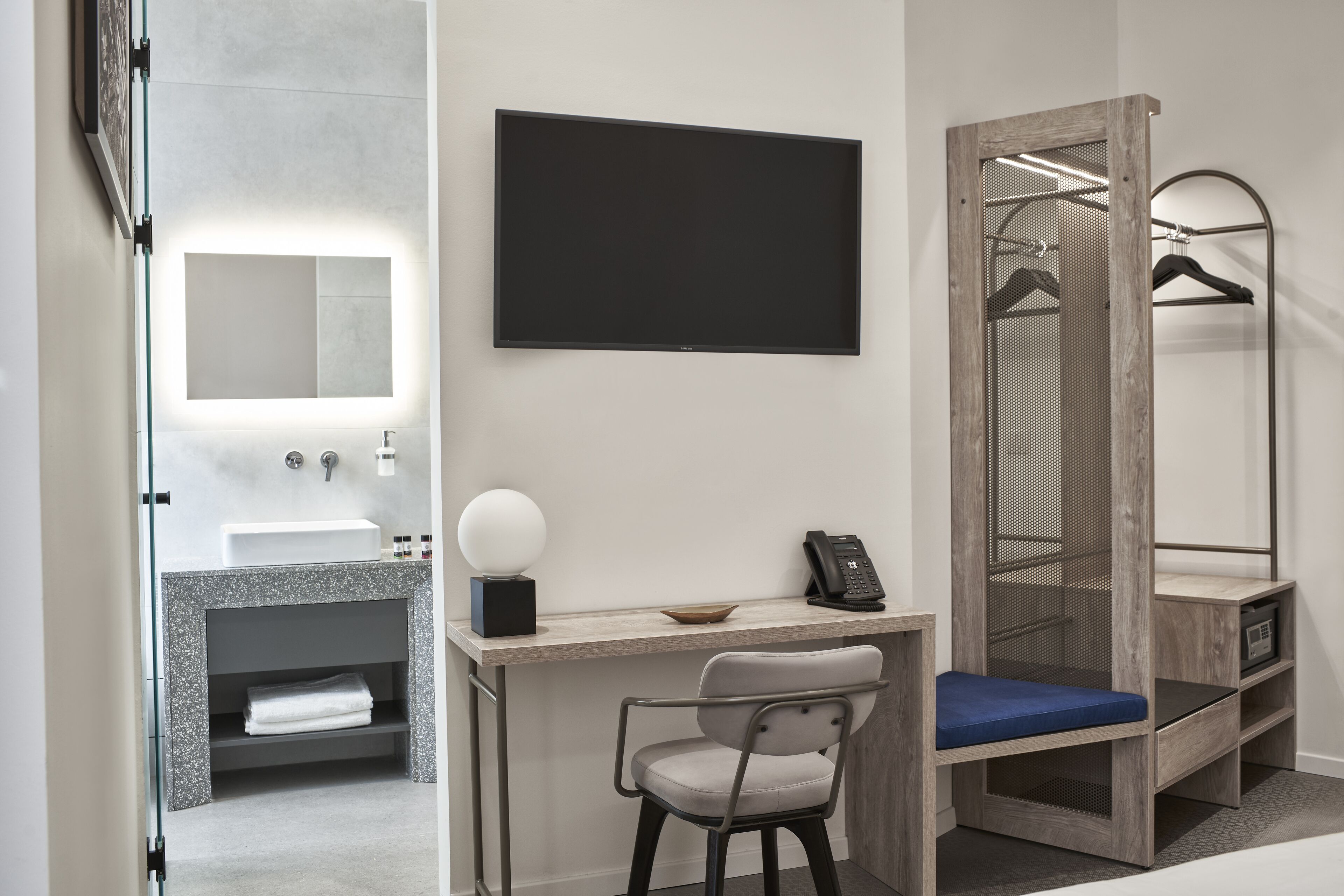 A modern hotel room with a large bed, a desk with a chair, a wardrobe, and a bathroom with a sink and mirror.
