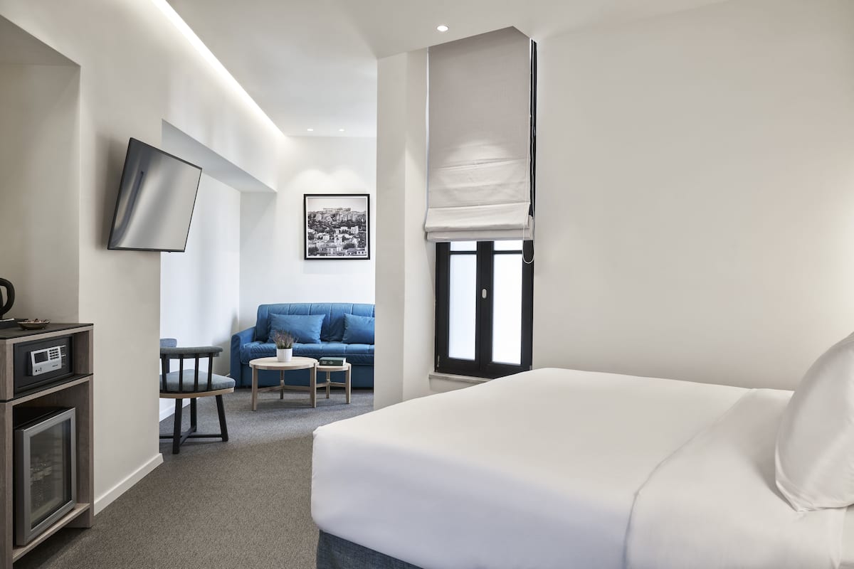 Habitación de hotel con una cama grande, un televisor en la pared, un sofá azul, una mesita pequeña y una ventana con persiana.