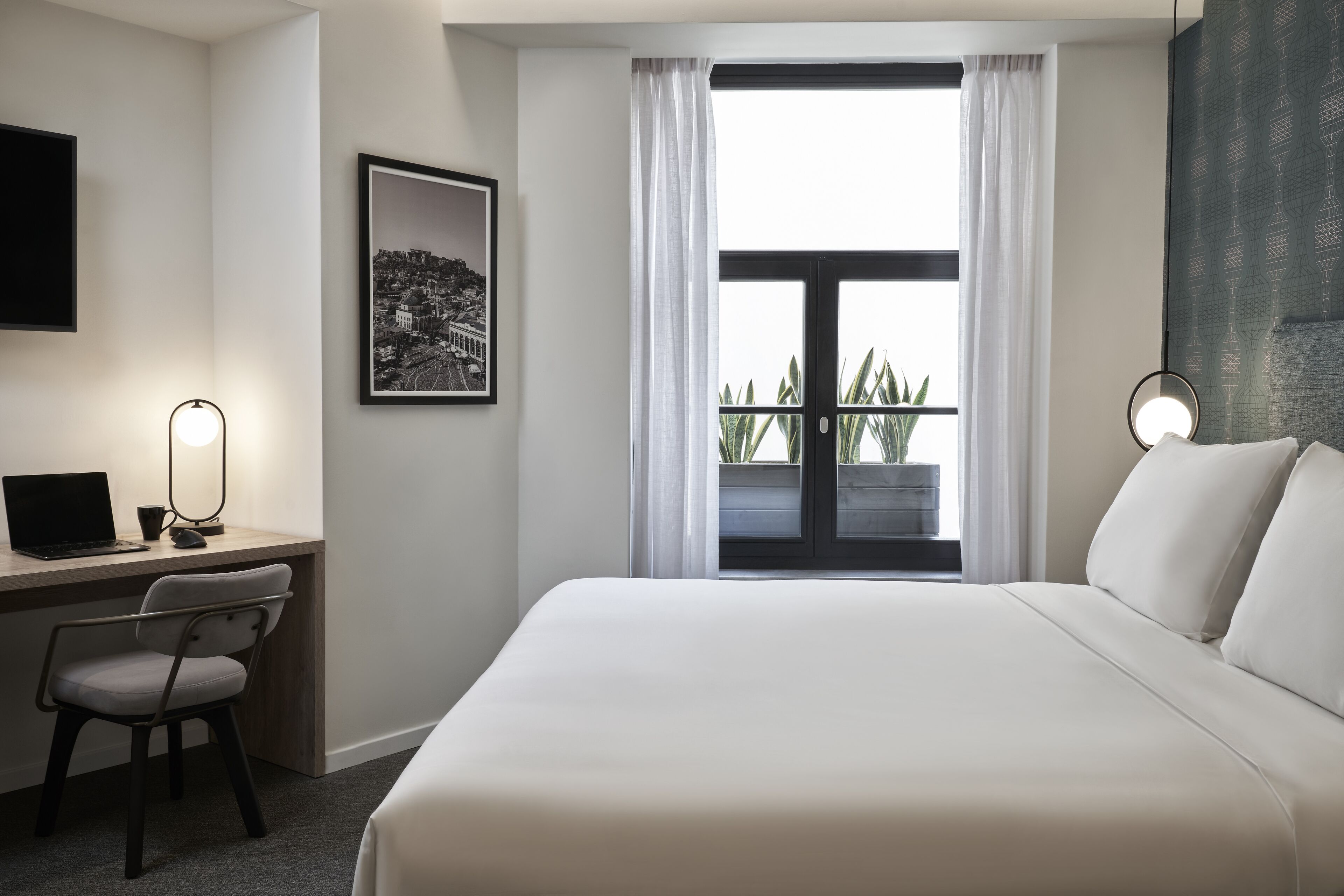 A hotel room with a bed, a desk with a laptop, a chair, a framed picture, and a window with curtains.