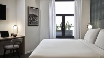 A hotel room with a bed, a desk with a laptop, a chair, a framed picture, and a window with curtains.