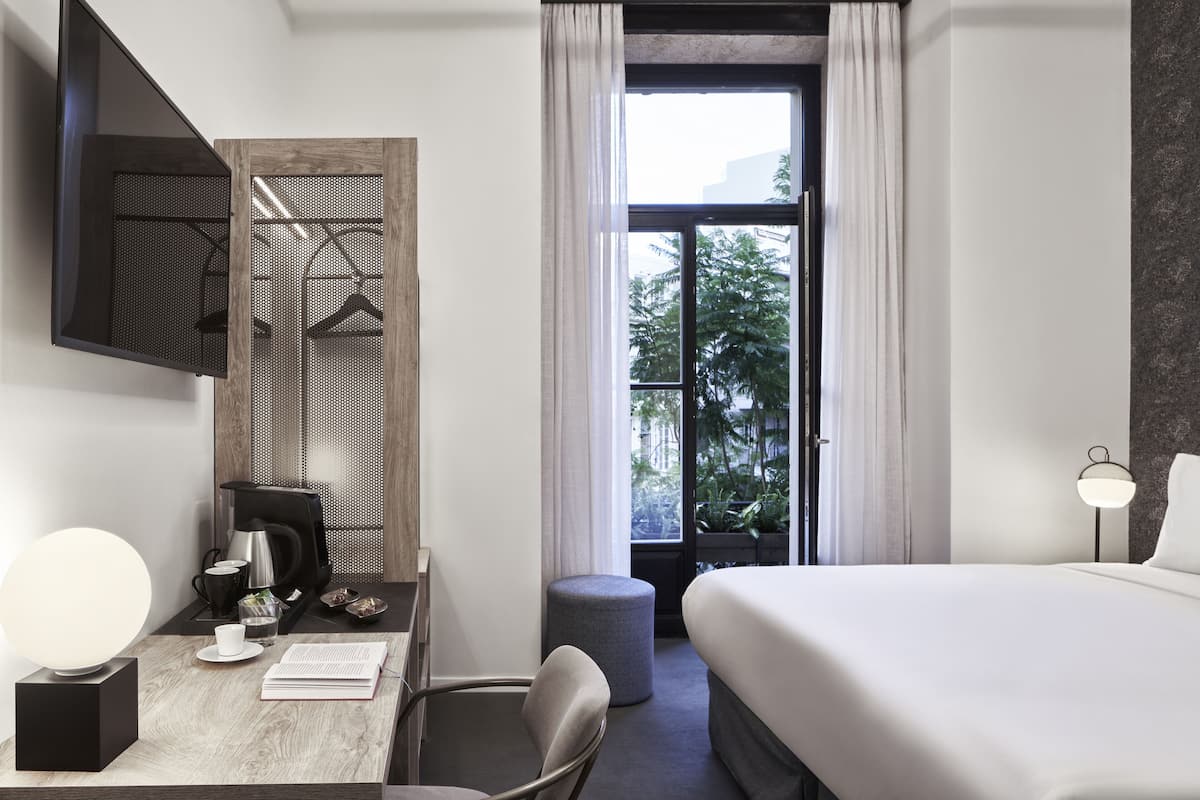 Una habitación de hotel moderna con una cama, un escritorio con cafetera, una silla, un reposapiés redondo pequeño, un armario con espejo y una ventana con vistas a zonas verdes.