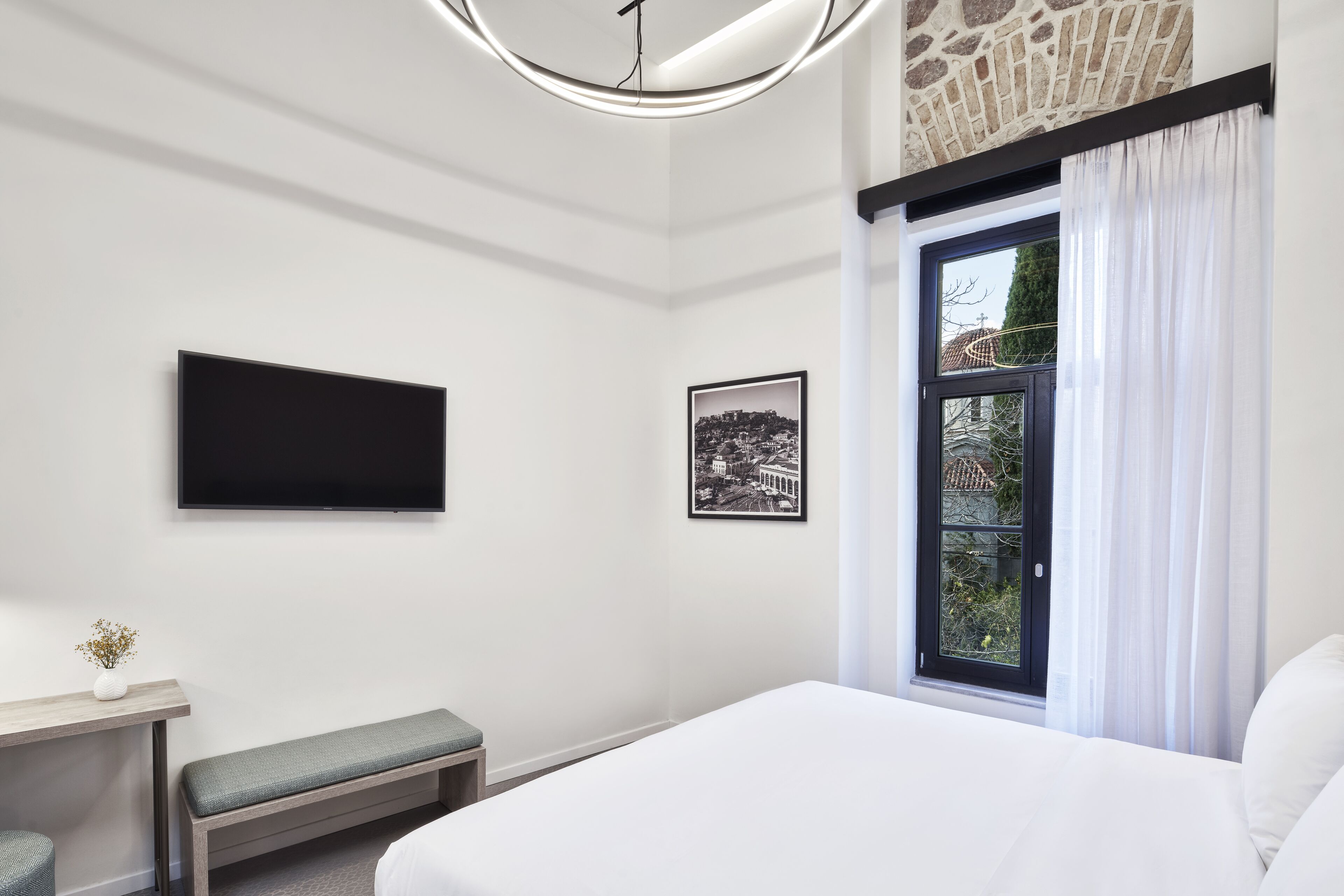 Una habitación de hotel moderna con una cama grande, un televisor de pantalla plana montado en la pared, un cuadro con una vista urbana enmarcada, un banco y una ventana con cortinas.