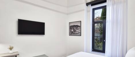 Una habitación de hotel moderna con una cama grande, un televisor de pantalla plana montado en la pared, un cuadro con una vista urbana enmarcada, un banco y una ventana con cortinas.
