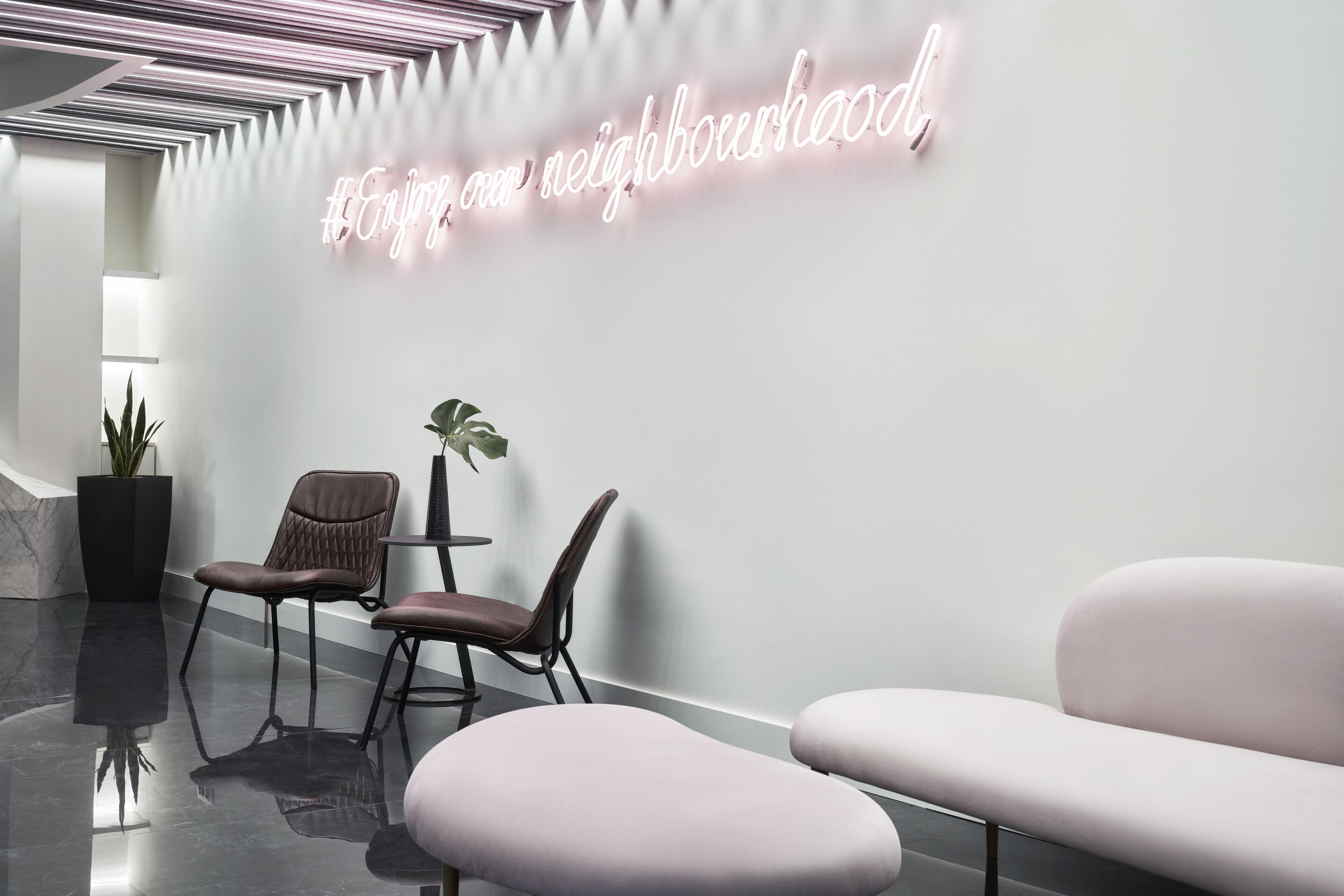 Un espace d'accueil moderne agrémenté d'un panneau lumineux néon, de deux coins salon avec des fauteuils confortables et d'une plante en pot.