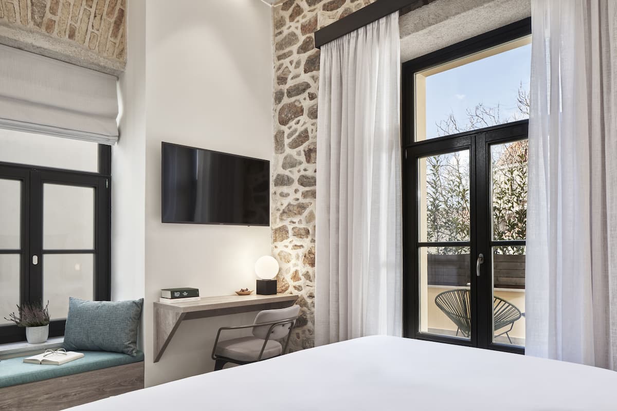 Un dormitorio con pared de piedra decorativa, un ventanal con cortinas, una mesita de noche con lámpara, una silla y un televisor montado en la pared.