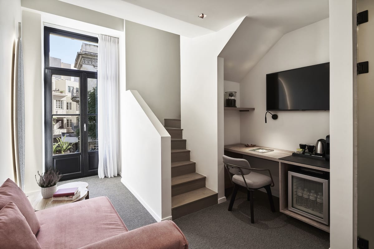 Un apartamento moderno con una escalera que lleva a un balcón, un escritorio pequeño con una silla, un televisor montado en la pared y una zona de estar acogedora con un sofá y una planta.