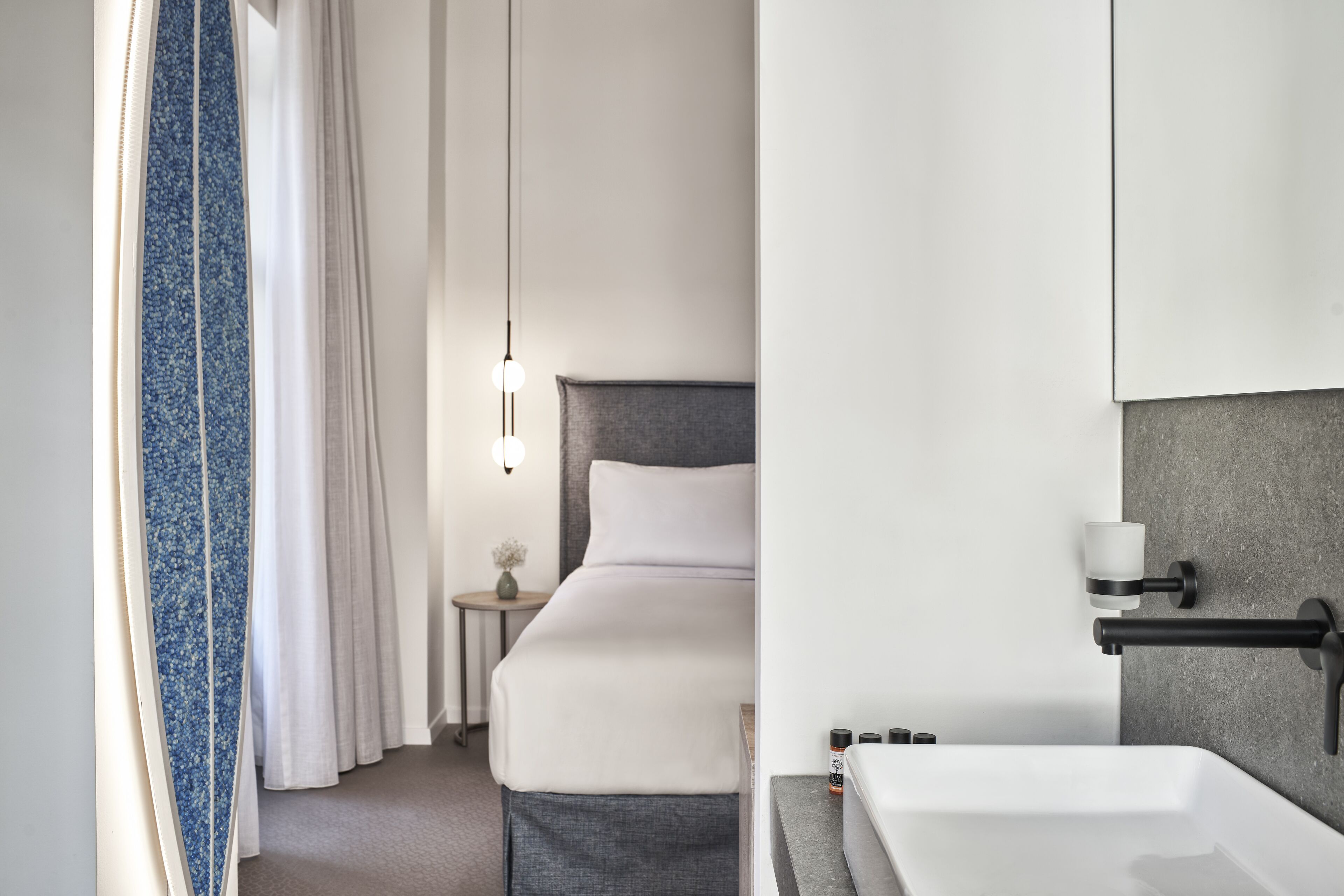 A modern hotel room with a large bed, a bedside table with a lamp, a bathroom with a sink and mirror, and a shower area.