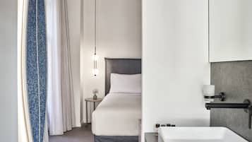 A modern hotel room with a large bed, a bedside table with a lamp, a bathroom with a sink and mirror, and a shower area.