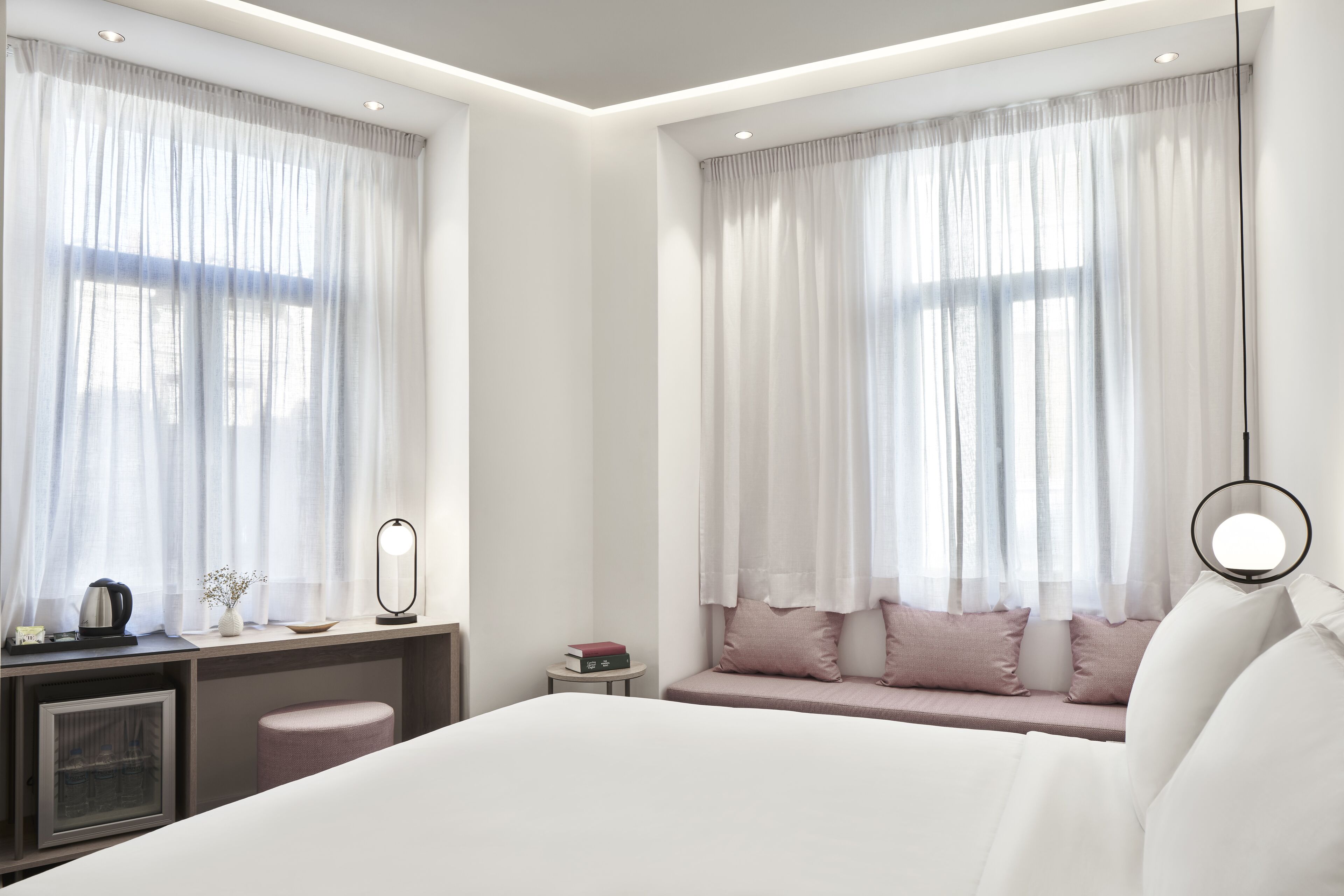 A modern hotel room with a large bed, a desk with a mirror, a small table, and a seating area with cushions.
