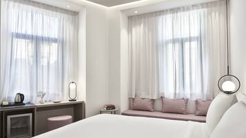 A modern hotel room with a large bed, a desk with a mirror, a small table, and a seating area with cushions.