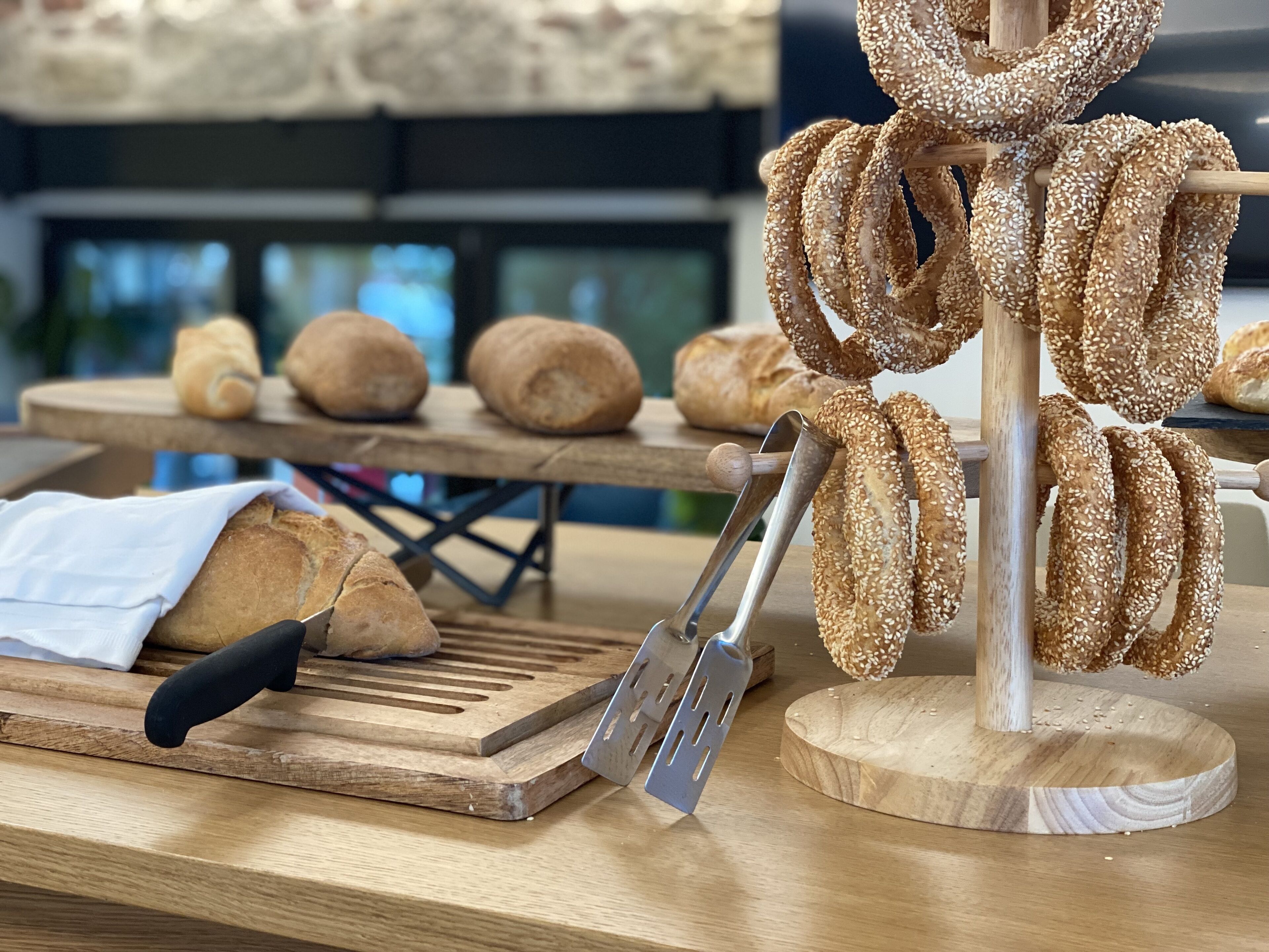 Une planche à découper en bois, accompagnée d’une miche de pain, d’un tranche-pain et d’une sélection de petits pains aux graines de sésame.