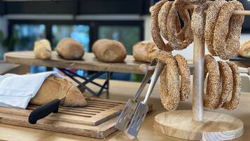 Une planche à découper en bois, accompagnée d’une miche de pain, d’un tranche-pain et d’une sélection de petits pains aux graines de sésame.