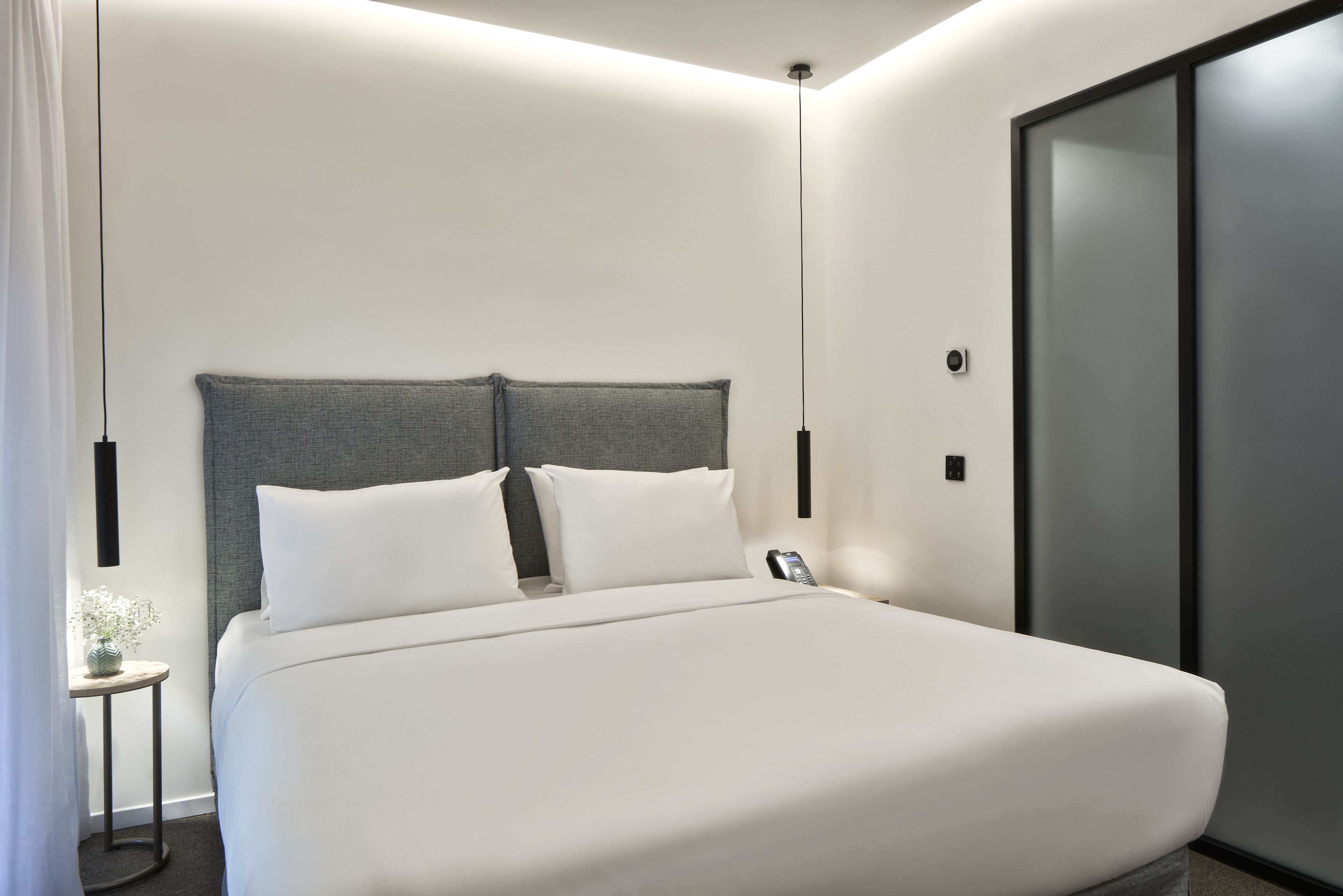 Una habitación de hotel moderna con una cama grande, una mesita de noche y un pequeño jarrón con flores.
