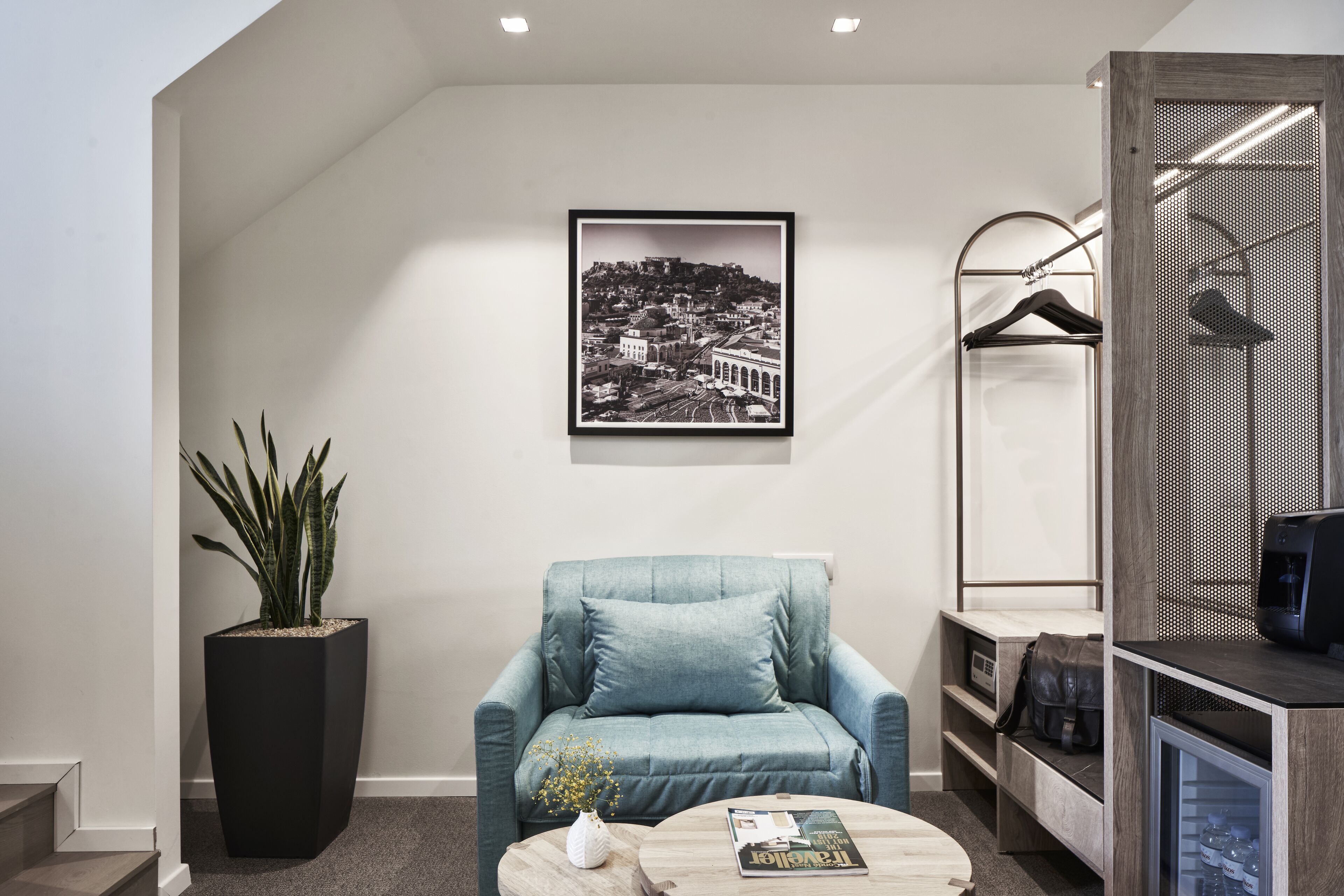 A modern room with a large potted plant, a blue armchair, a coffee table with a magazine, a framed cityscape photo, and a wardrobe with a hanging rack.