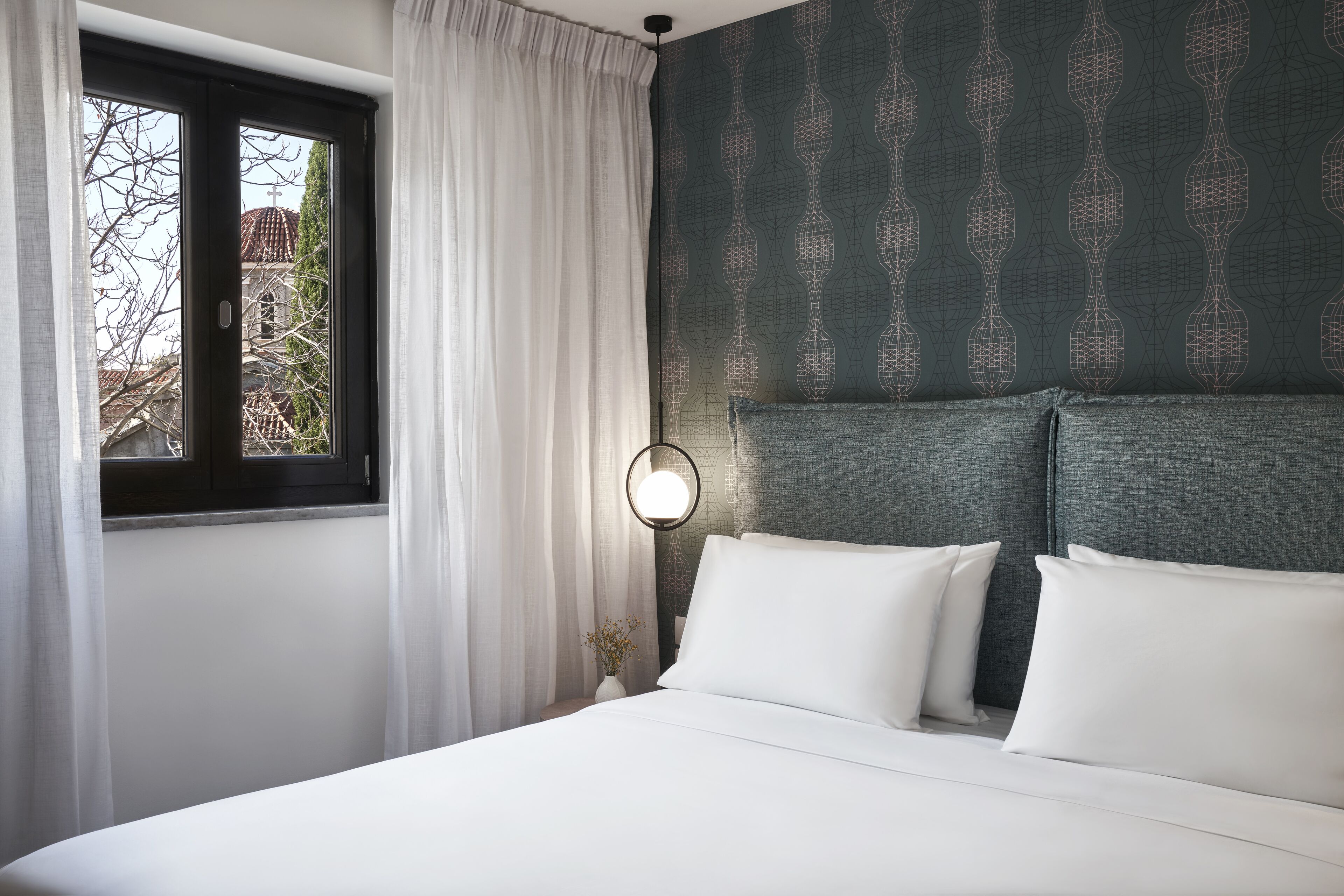 A neatly made bed with white linens, a bedside lamp, and a window with sheer curtains.