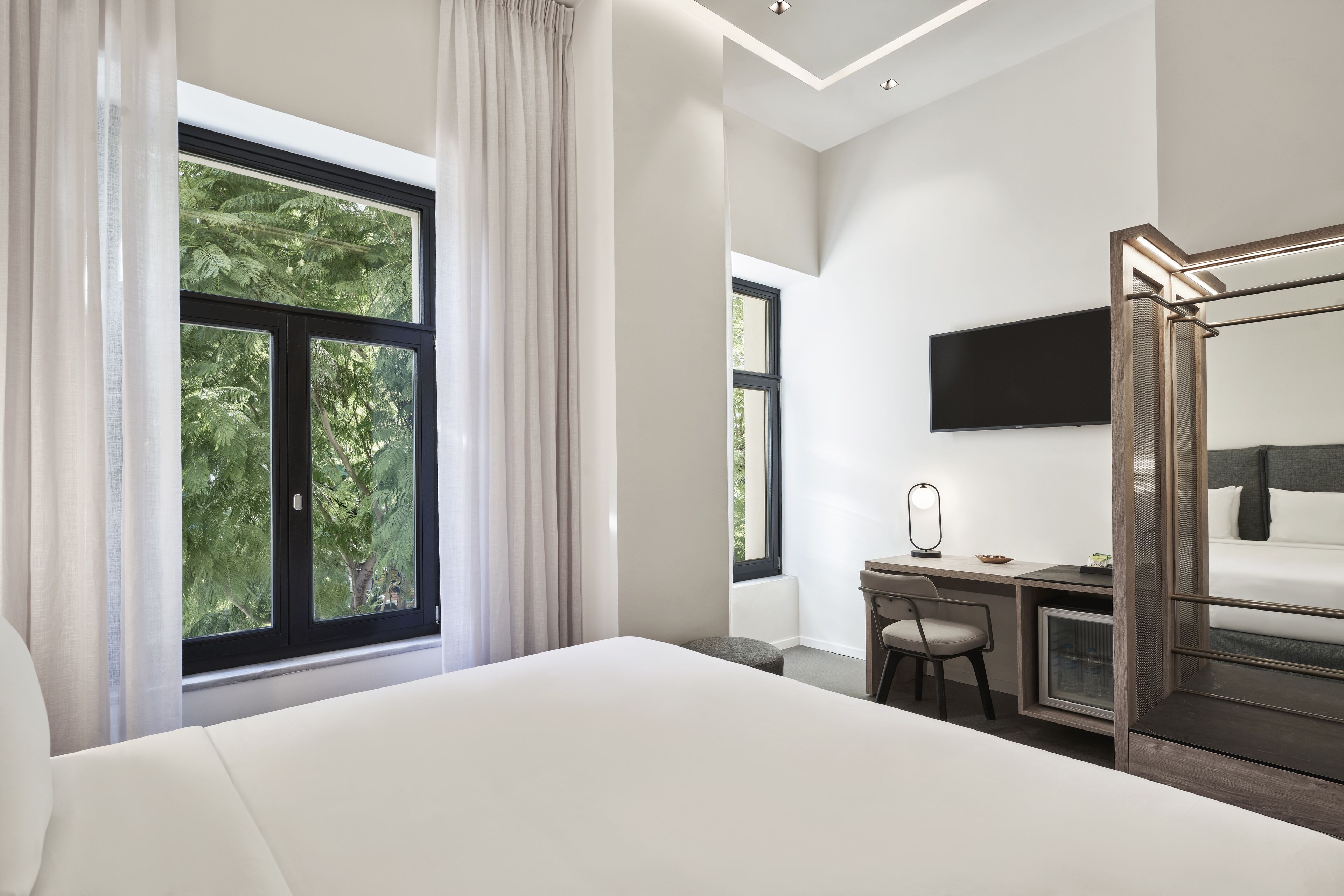 A modern hotel room with a large bed, a desk with a chair, a TV mounted on the wall, and a window with curtains.