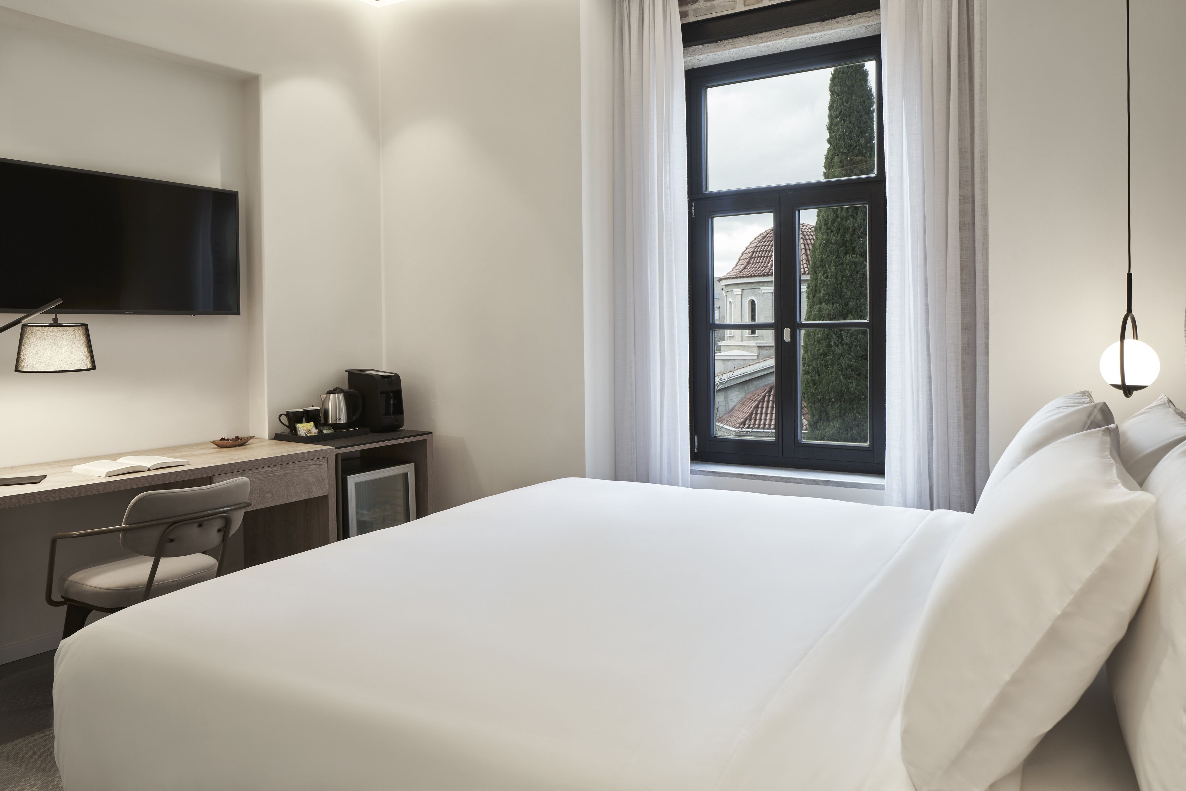 A modern hotel room with a large bed, a desk with a chair, a flat-screen TV, and a window with a view of a building.