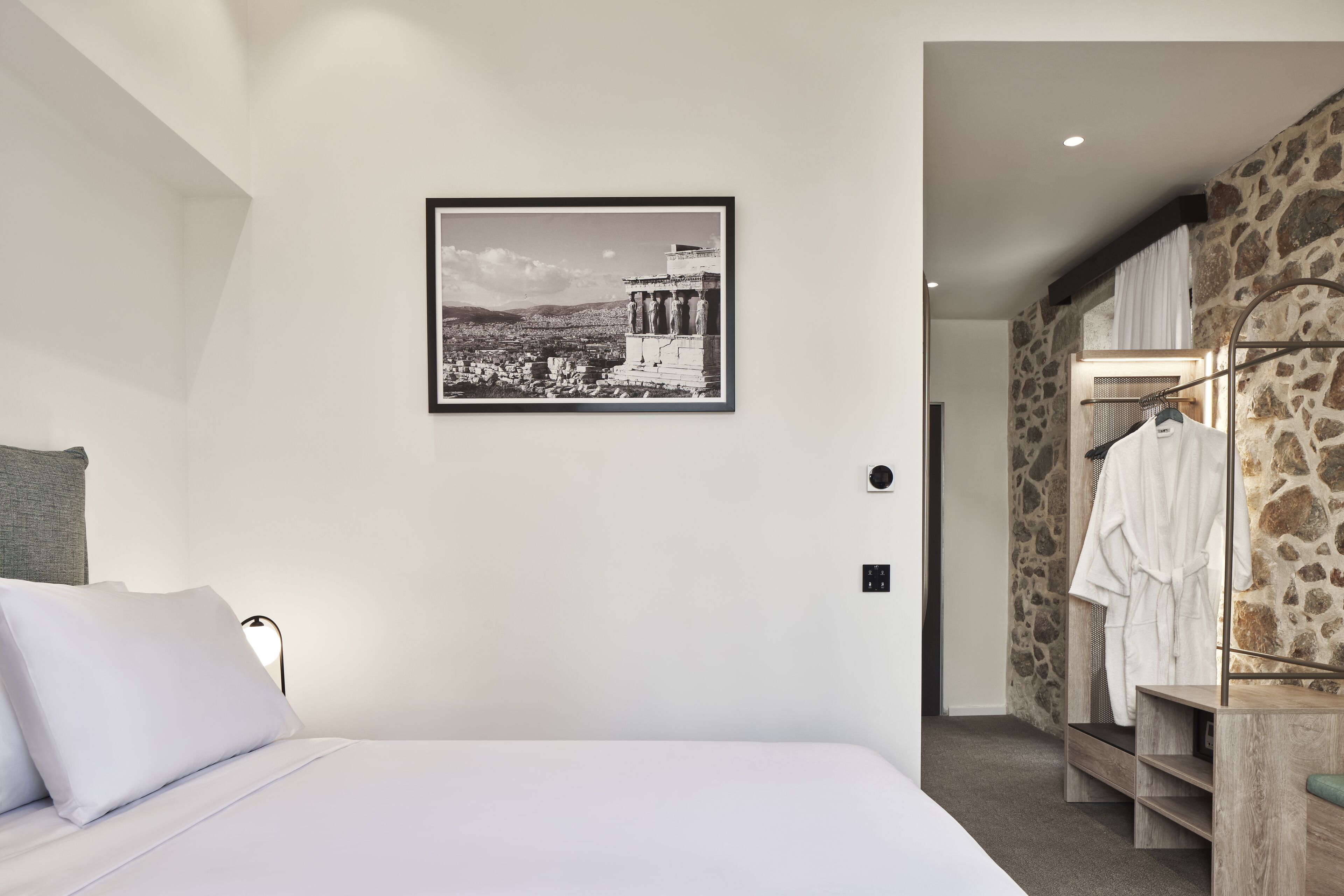 A bedroom with a bed, a framed picture of a historical building, a closet with a robe, and a stone wall.