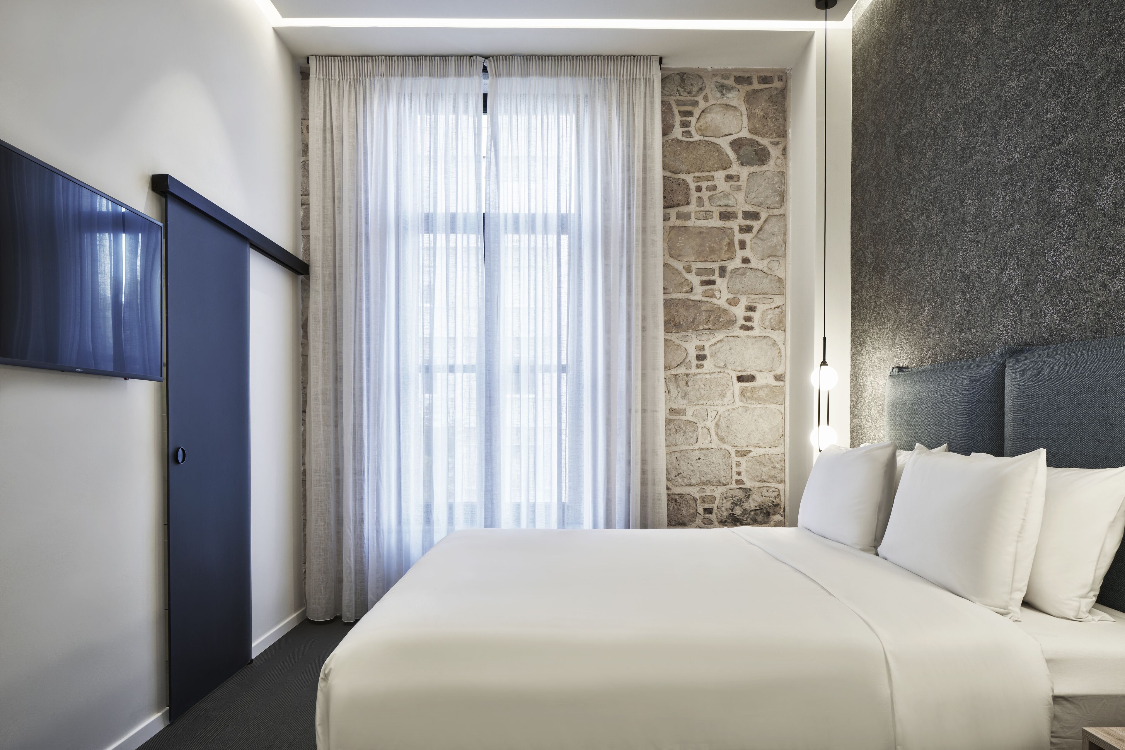 A hotel room with a large bed, a stone accent wall, a dark headboard, and a large window with sheer curtains.