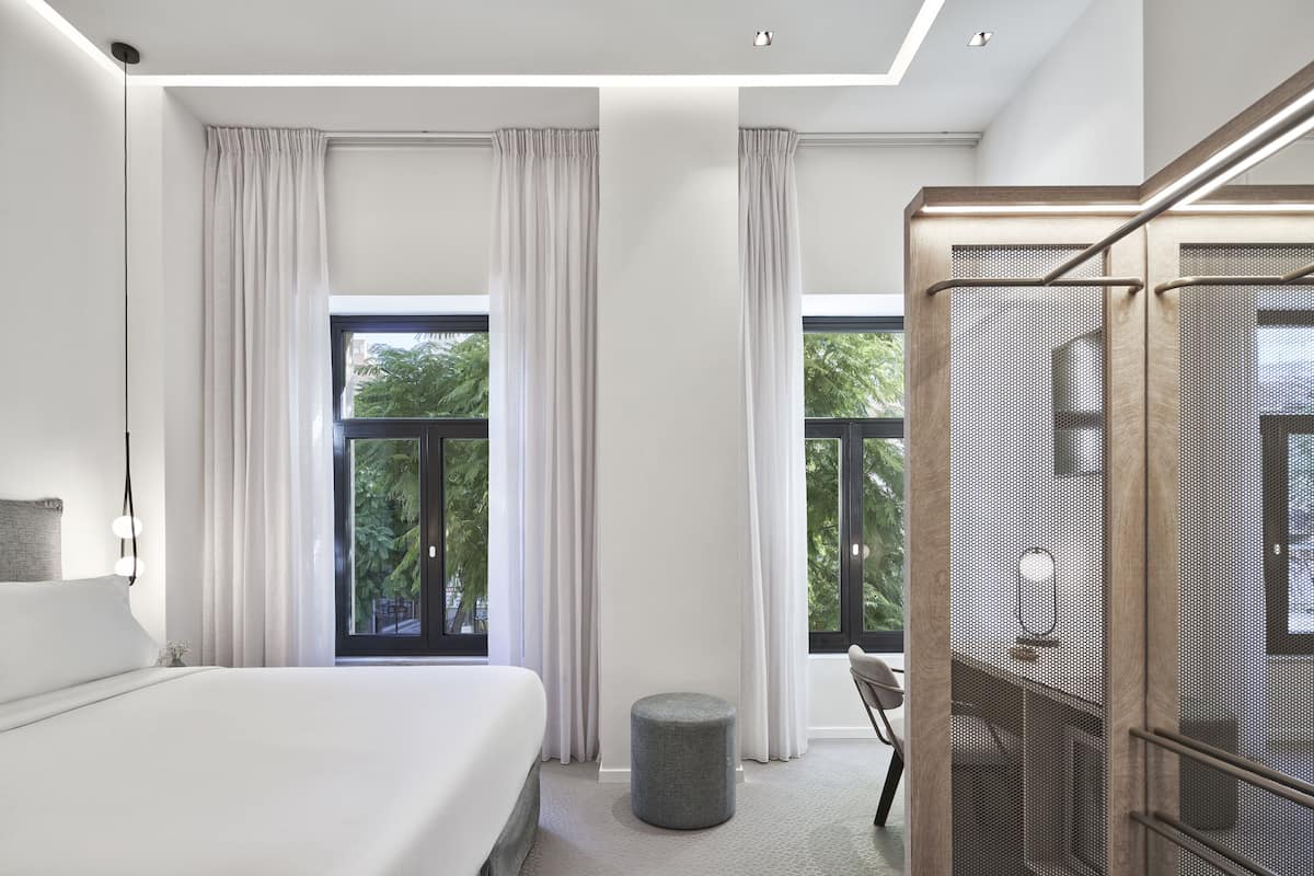 Una habitación de hotel moderna con una cama grande, un otomanillo redondo y un vestidor con espejo y ropa colgada.