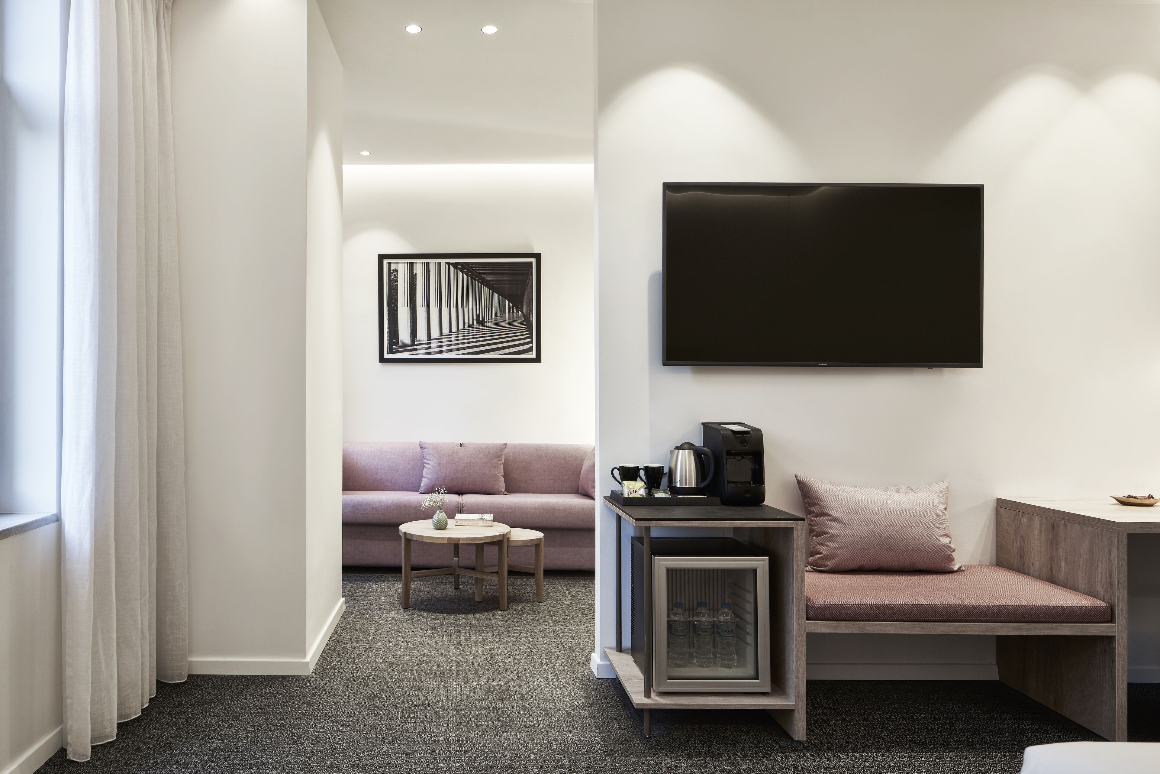 A modern hotel room with a sofa, a small table, a coffee machine, and a TV mounted on the wall.