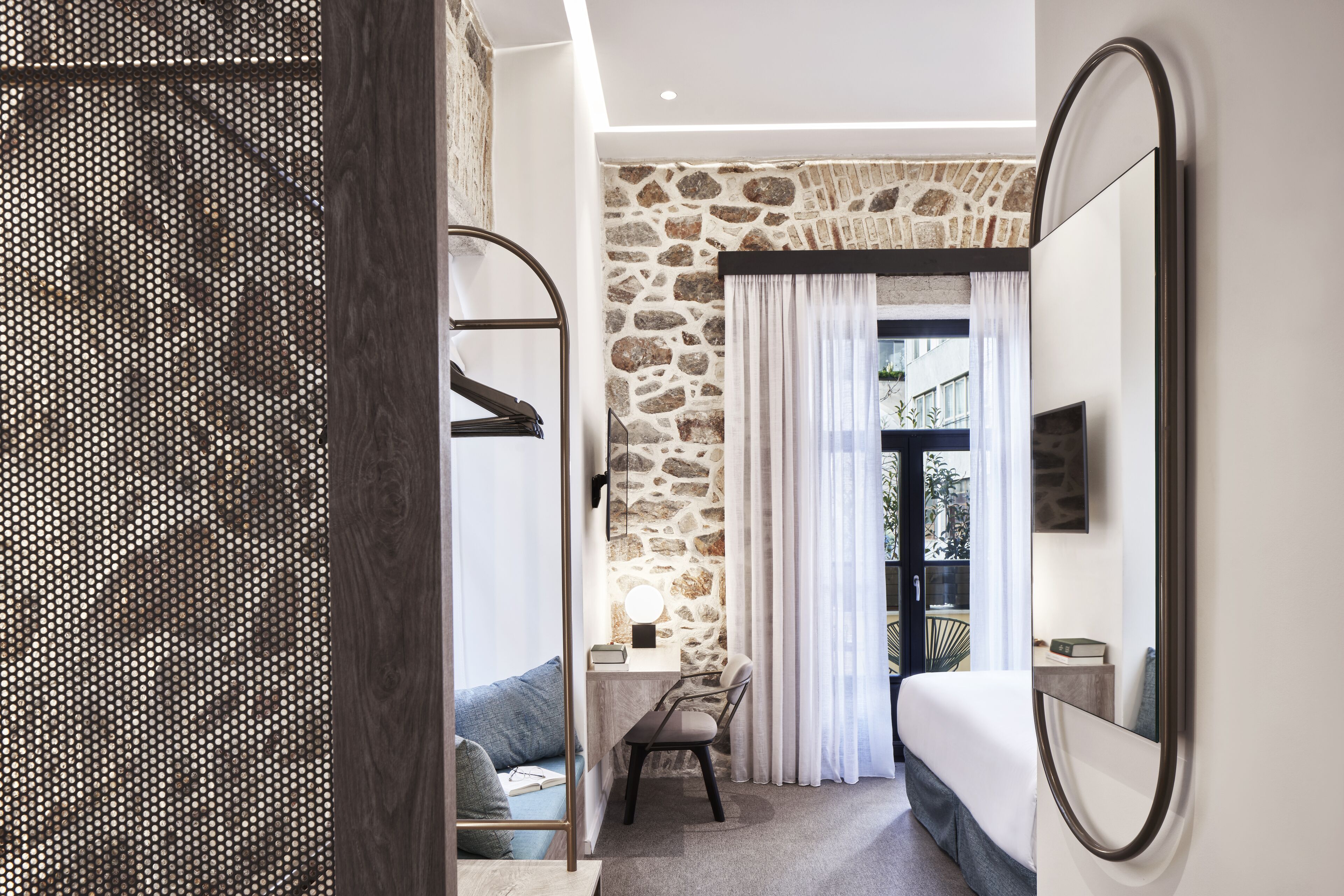 A modern hotel room with a stone accent wall, a desk with a chair, a bed, and a large mirror.