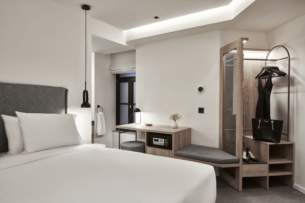 Una habitación de hotel moderna con una cama grande, un escritorio con una silla, un tocador con espejo y un perchero.