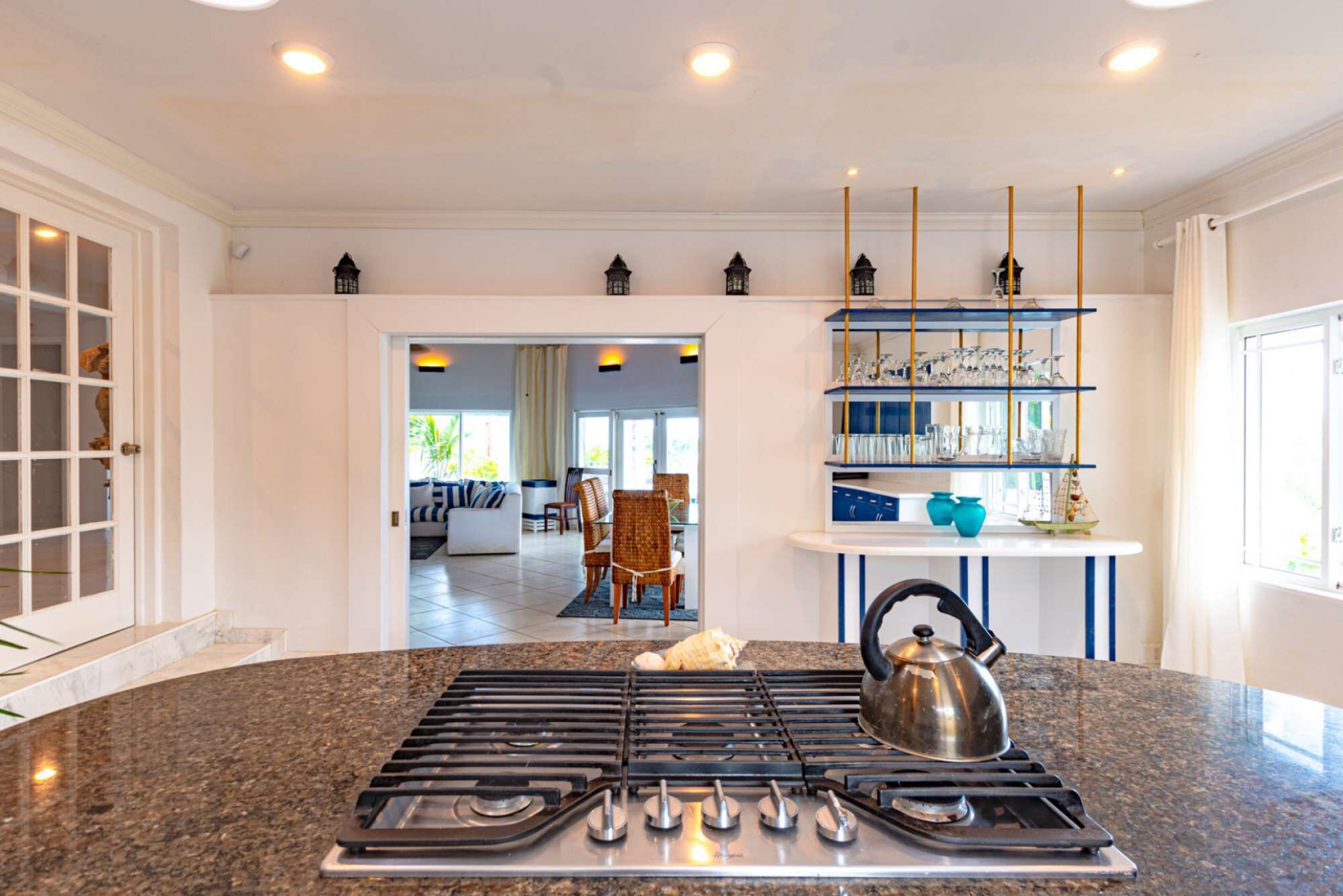 A kitchen with a gas stove, a kettle, and a shelf with glassware.