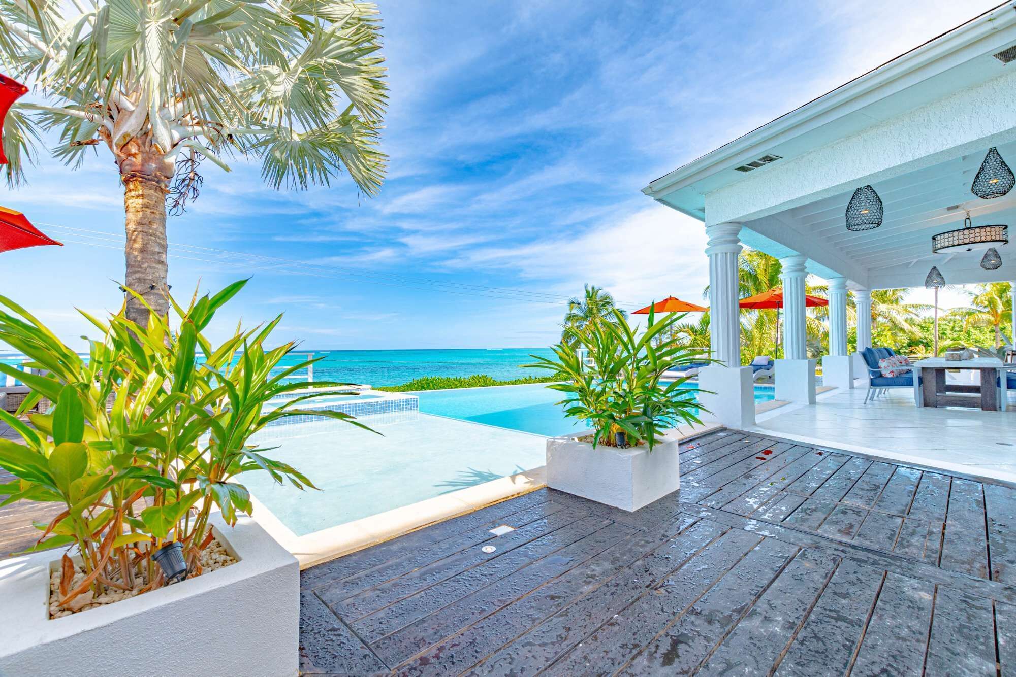 A poolside area with a view of the ocean, a covered seating area, and potted plants.