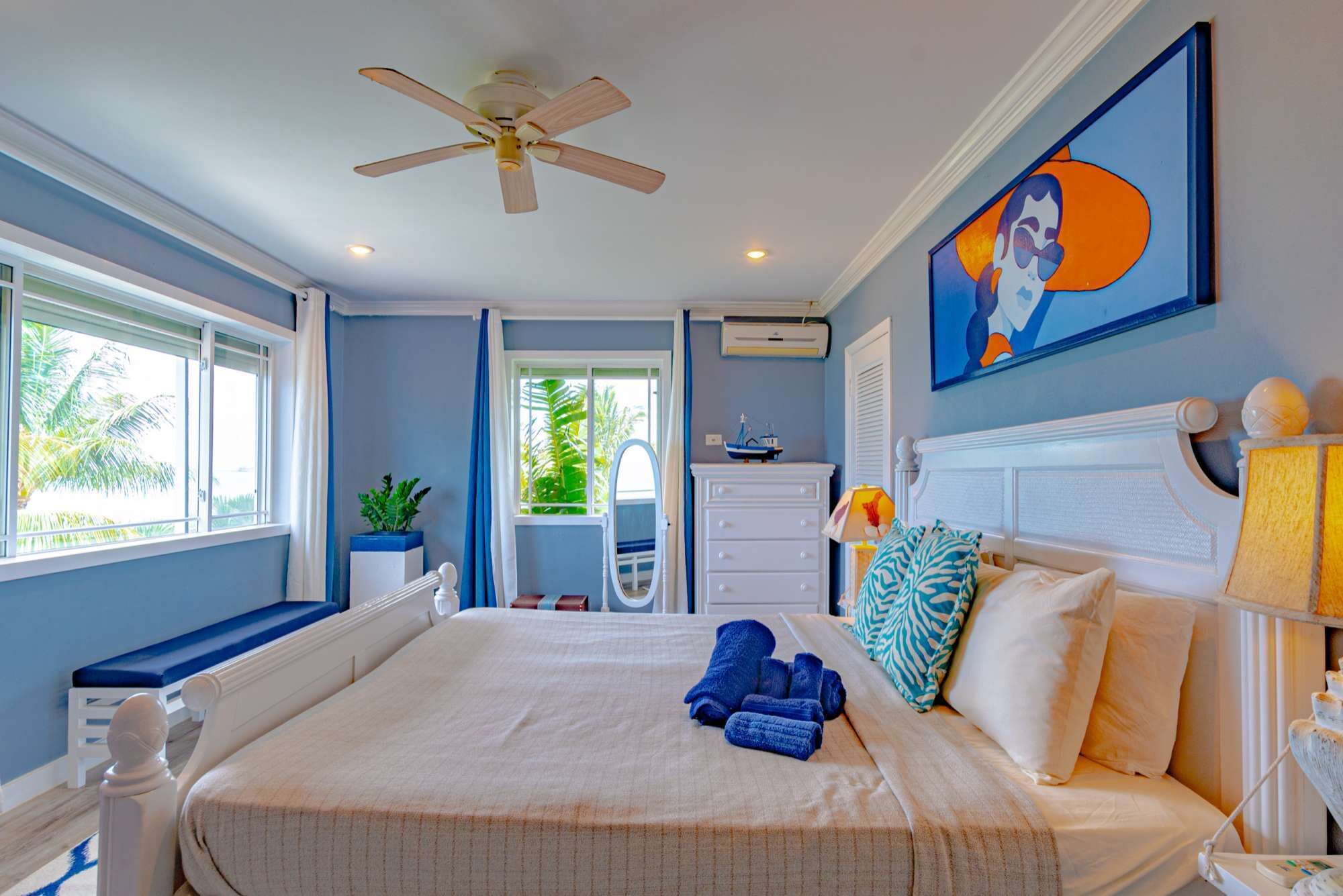 Een slaapkamer met een bed, een plafondventilator, een raam, een kamerplant, een dressoir en een schilderij aan de muur.