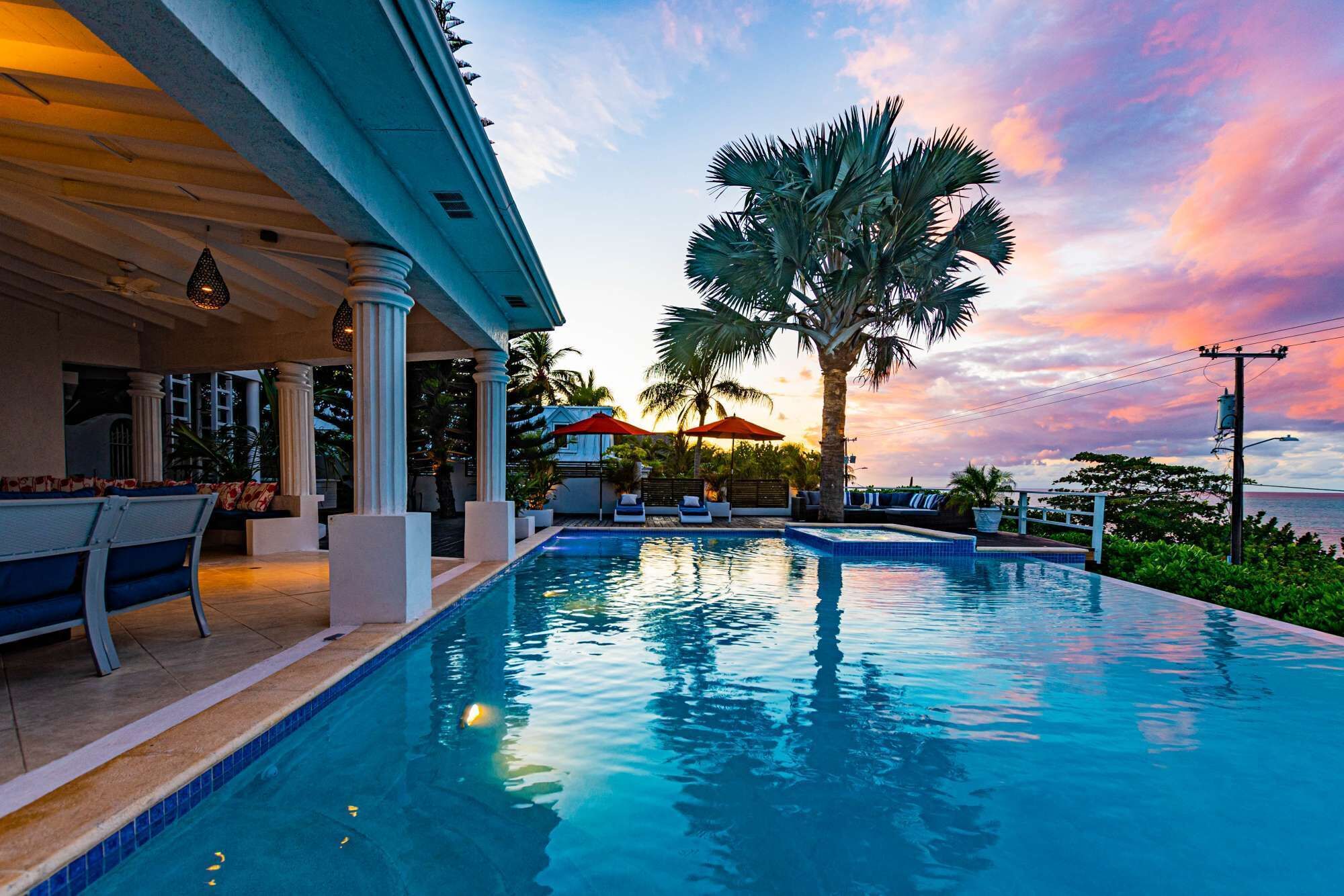 A swimming pool with lounge chairs and umbrellas, a palm tree, and a sunset view.