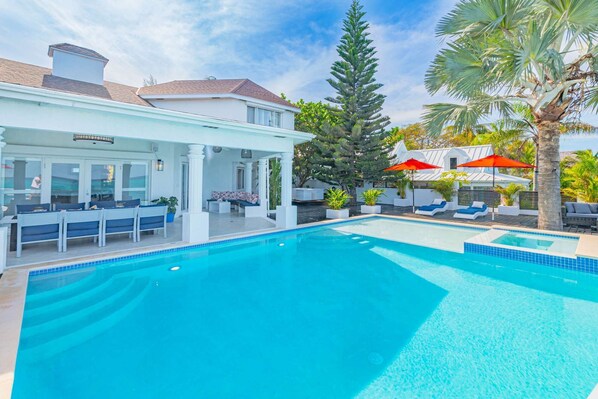 Ein Pool mit Liegestühlen und Sonnenschirmen, umgeben von Palmen und einer überdachten Sitzgruppe.