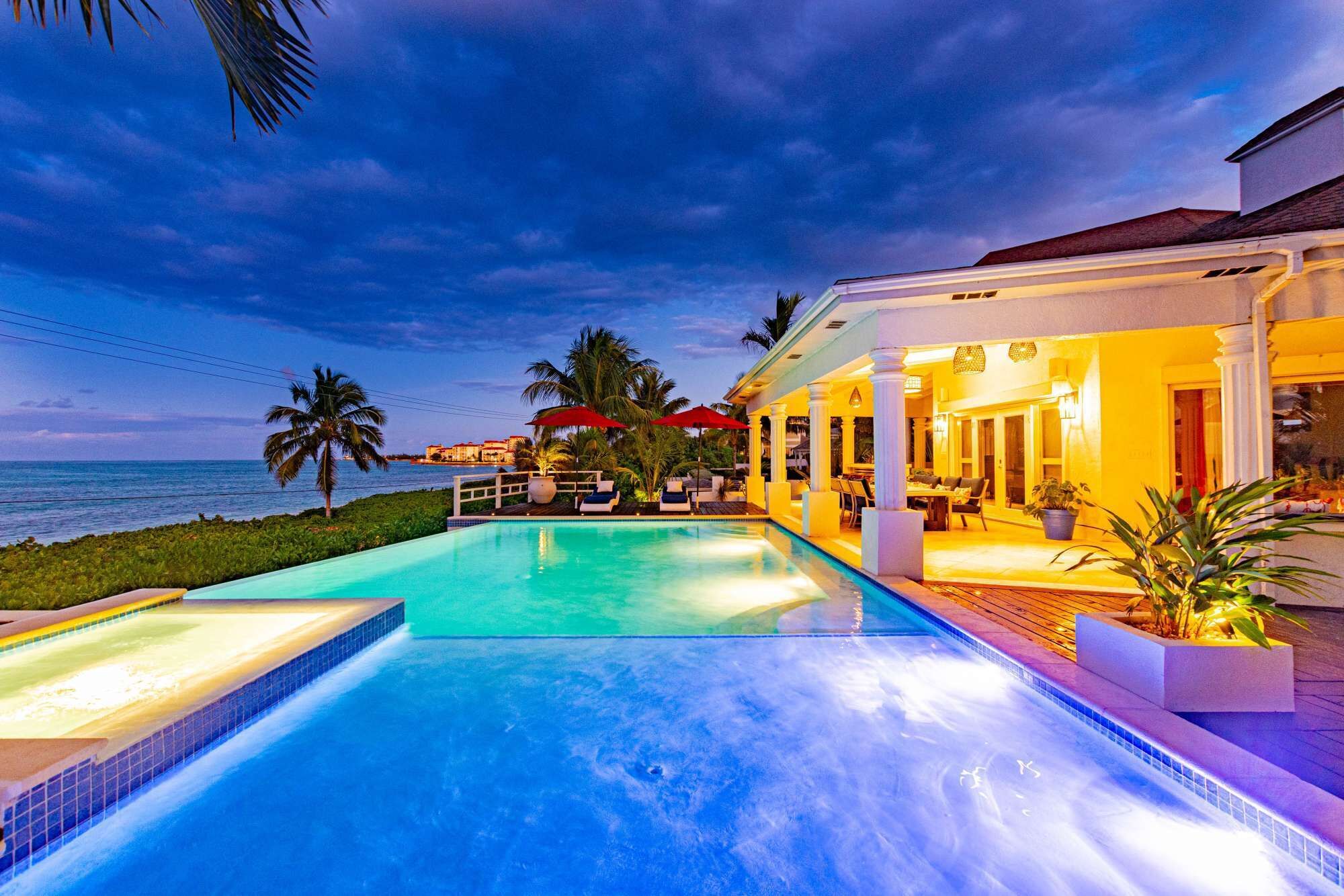 A swimming pool with a view of the ocean, a covered outdoor seating area, and a building with columns.