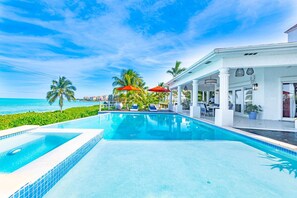 Ein Poolbereich mit Blick auf den Ozean, Palmen und ein Gebäude mit Außenbestuhlung.