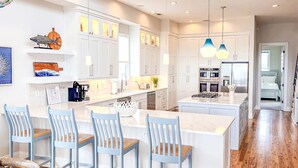 Een moderne keuken met witte kasten, een centraal eiland met barkrukken, hanglampen en uitzicht op de aangrenzende slaapkamer.