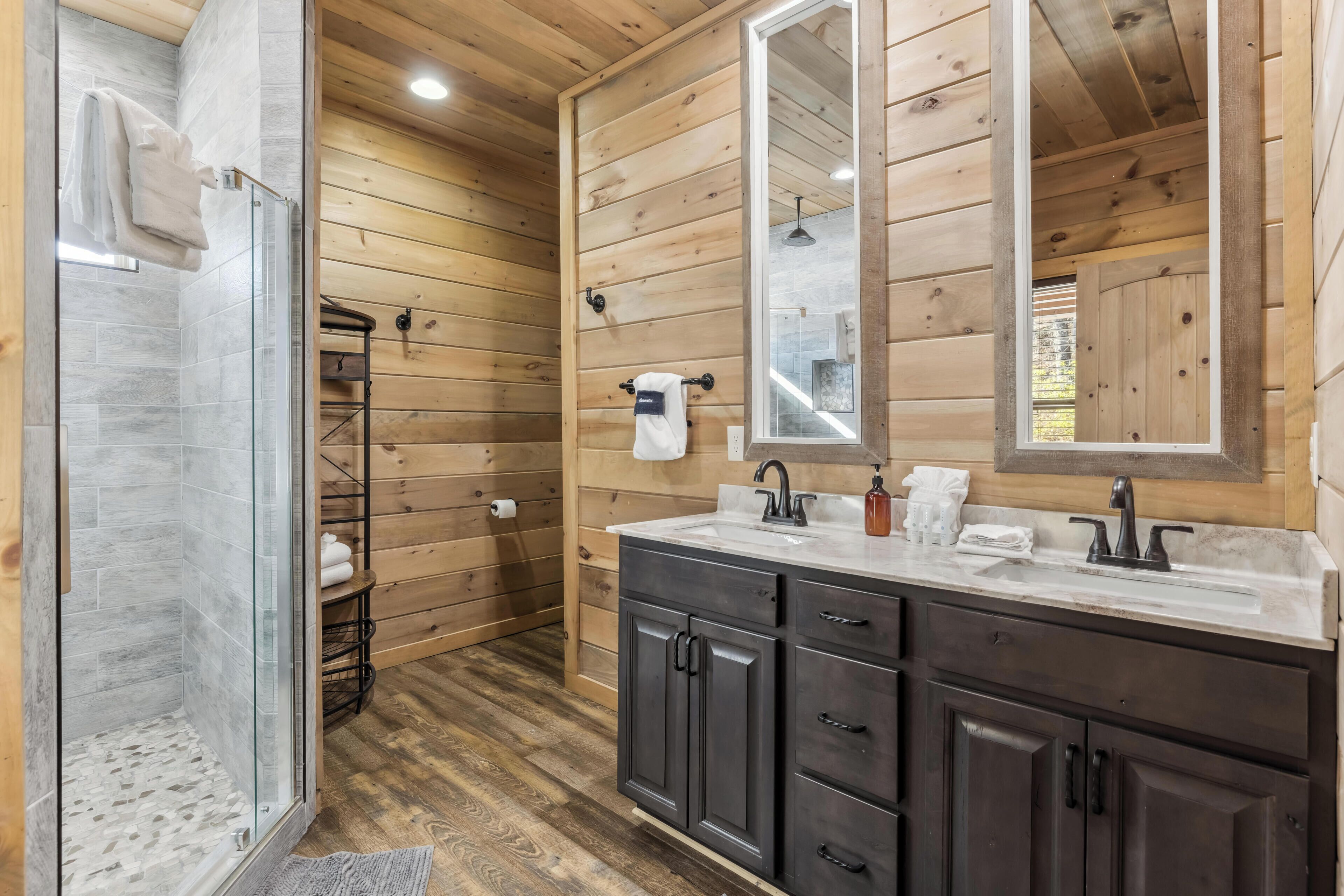 A wooden cabin bathroom with a glass shower, dark cabinets, and a large mirror.