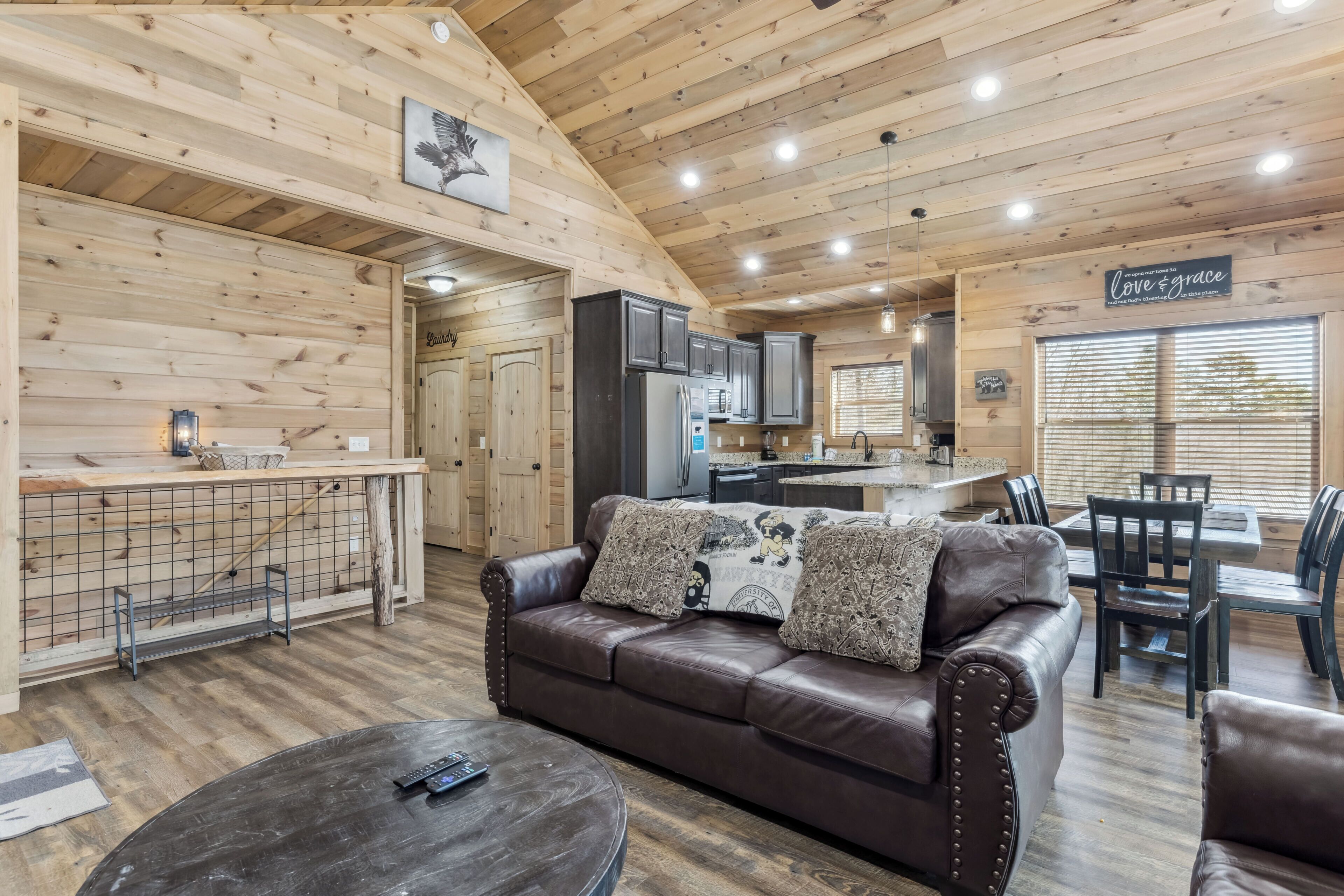 A cozy cabin interior with a wooden ceiling, a leather sofa, a dining area, and a kitchen.