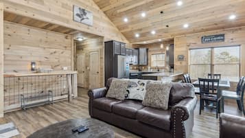 A cozy cabin interior with a wooden ceiling, a leather sofa, a dining area, and a kitchen.