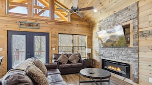 Interior acogedor de una cabaña con chimenea de piedra, sofás de cuero y un televisor grande montado en la pared.