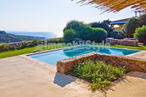 Una piscina con borde de piedra, rodeada de vegetación y vista al mar.