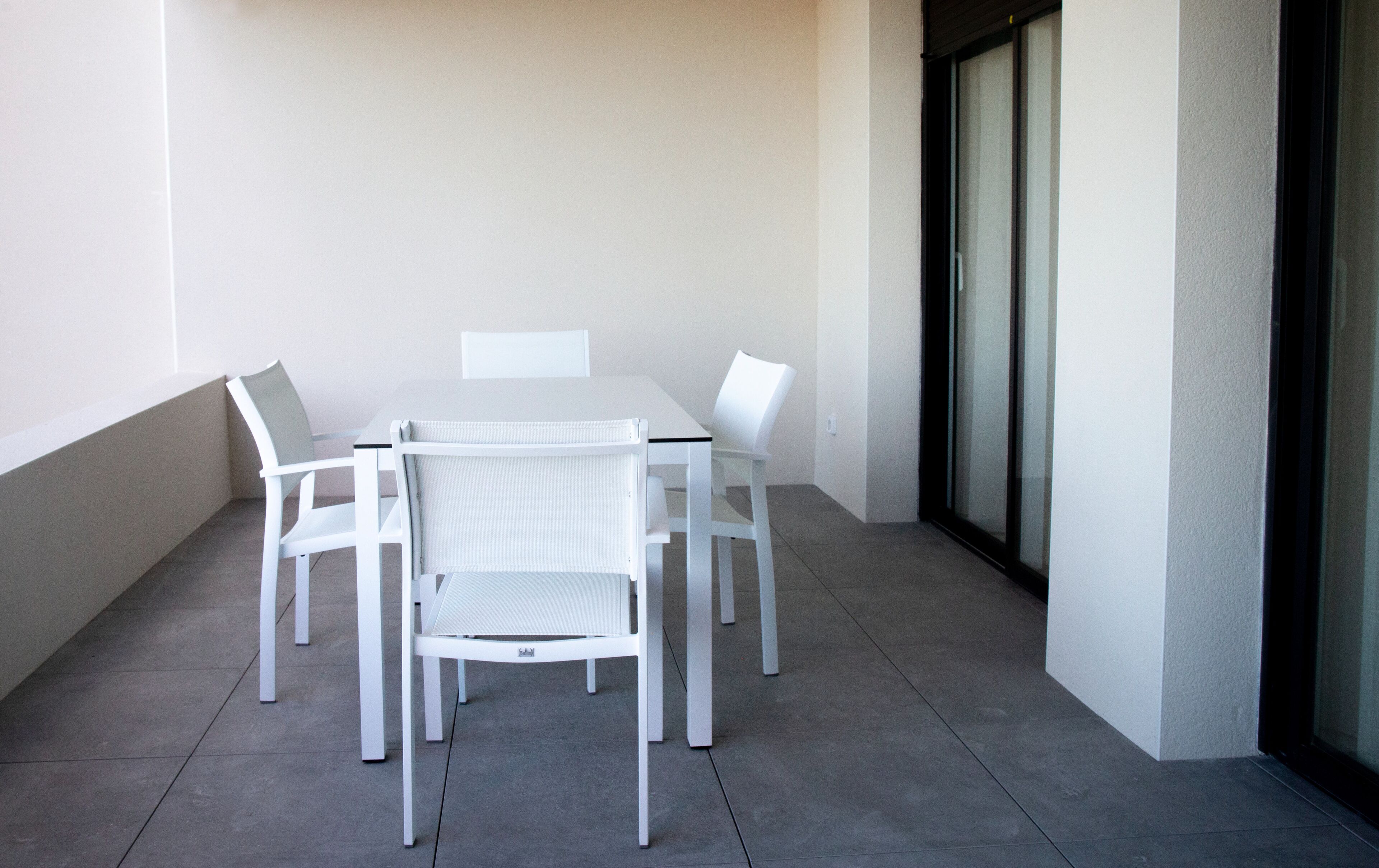 Une table blanche et quatre chaises sur un balcon doté d’un sol carrelé et d’une porte vitrée.