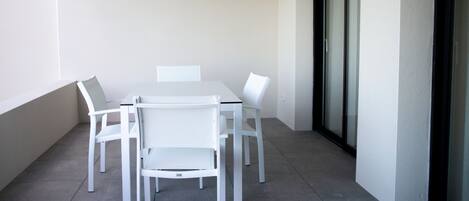 Una mesa blanca y cuatro sillas en un balcón con piso de baldosas y puerta de vidrio.